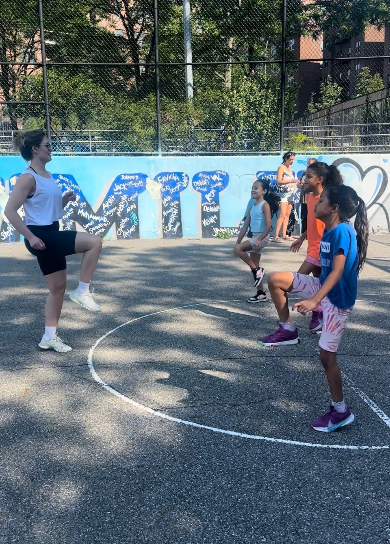 A group of children and a woman on a basketball court participating in a fitness class or exercise session, with a colorful mural and trees in the background.