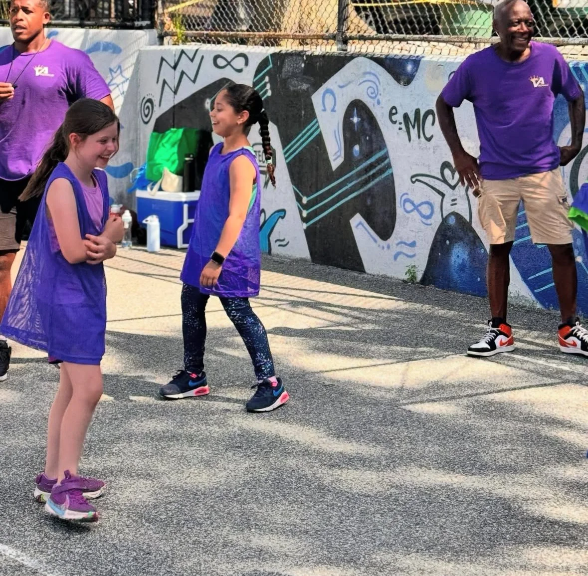 Youth girl athletes smiling and laughing, on a basketball court with graffiti art on the wall behind them, while coaches in purple shirts stand nearby.