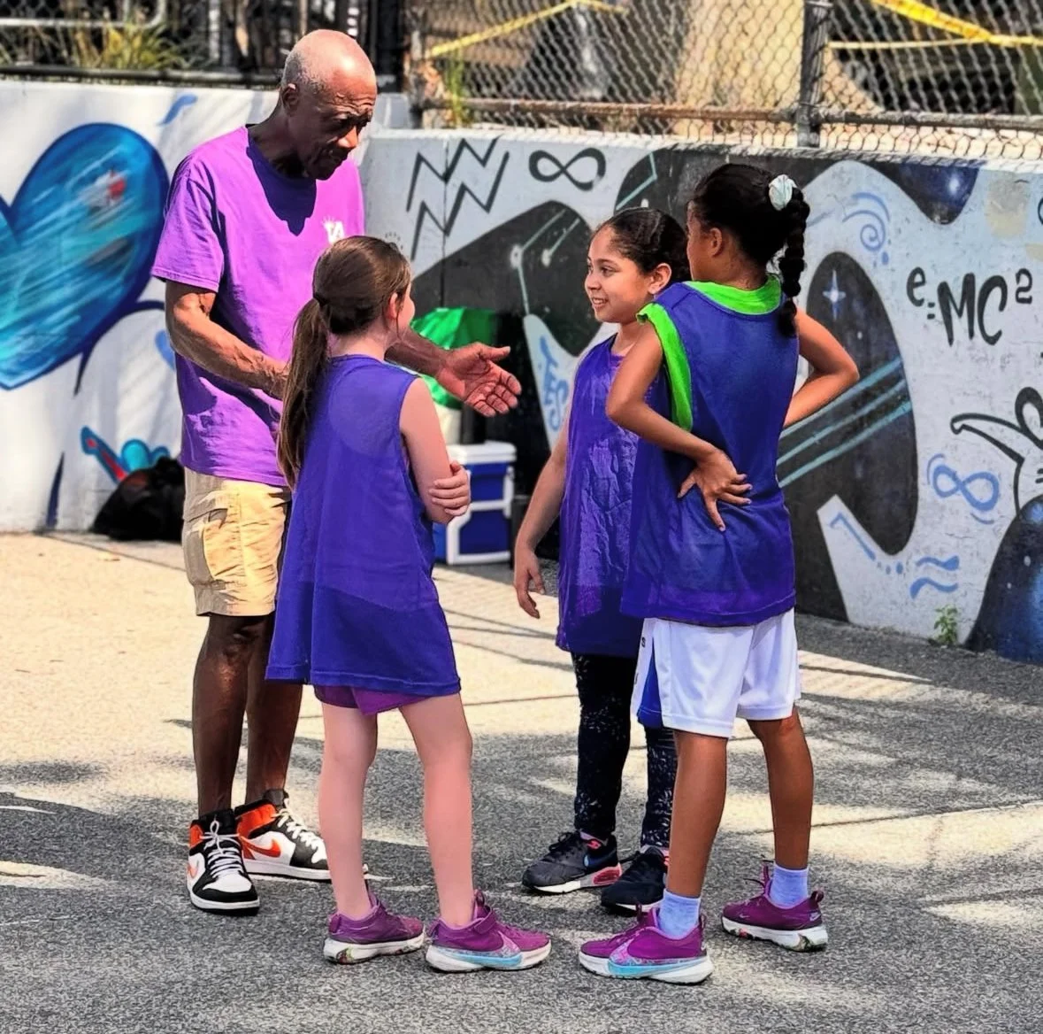 A coach or adult talking to three young girls on a basketball court, with a graffiti wall in the background and the girls wearing blue basketball vests.