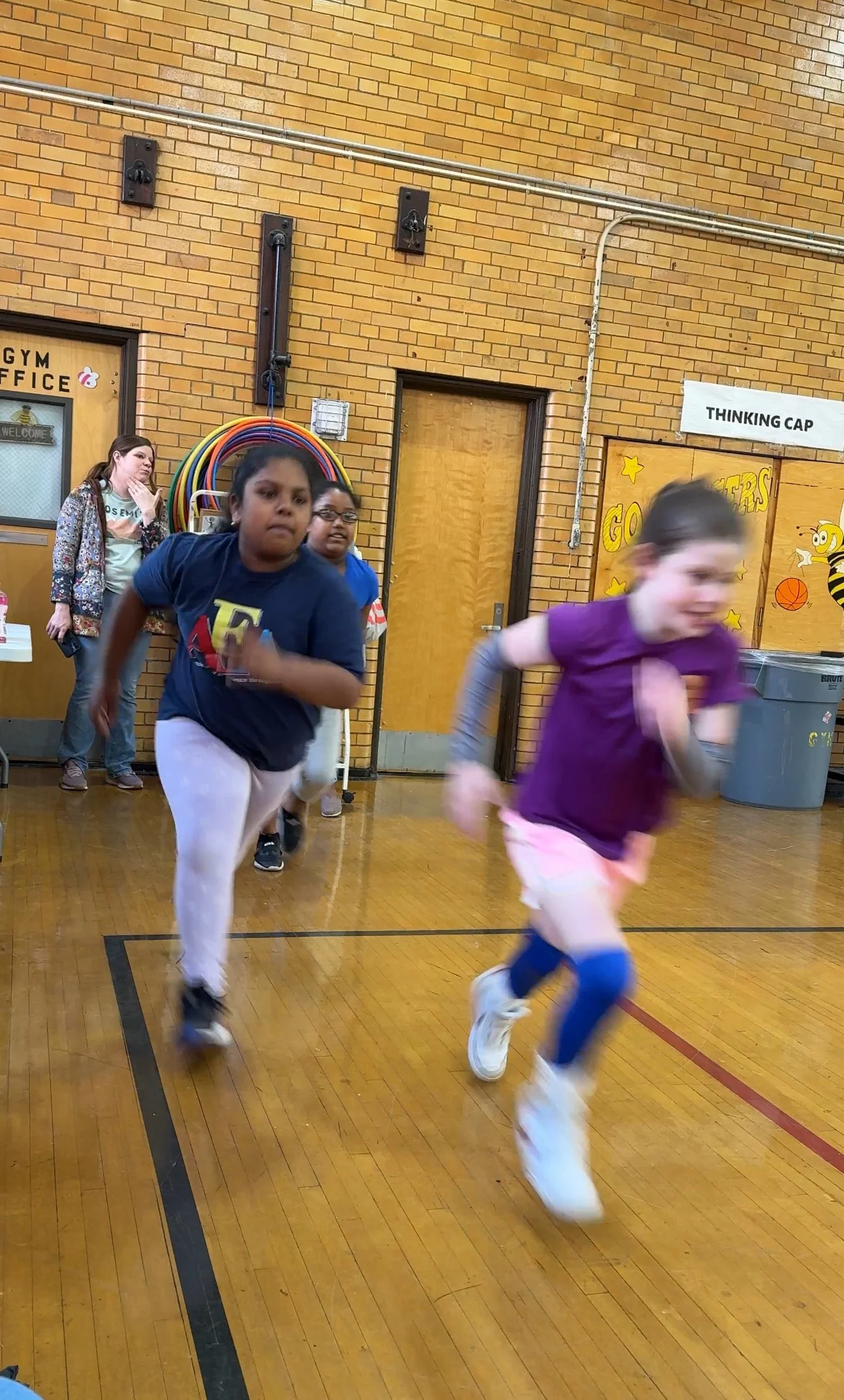 Two girls sprinting in a gymnasium, with other children with colorful signs and decorations.