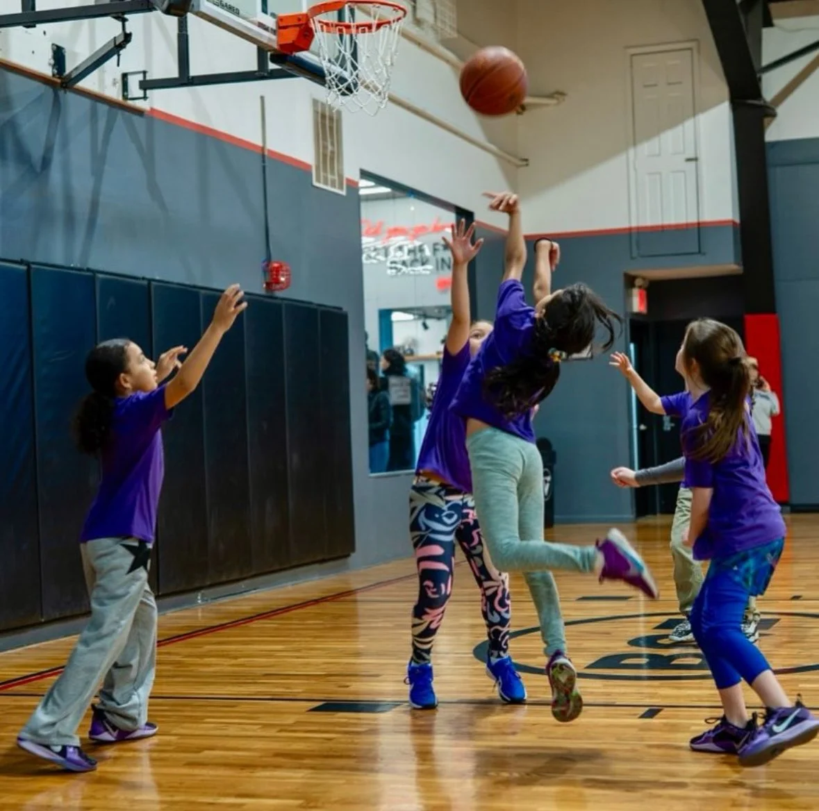 Children playing basketball in a gym, with one girl jumping to shoot the ball towards the hoop.