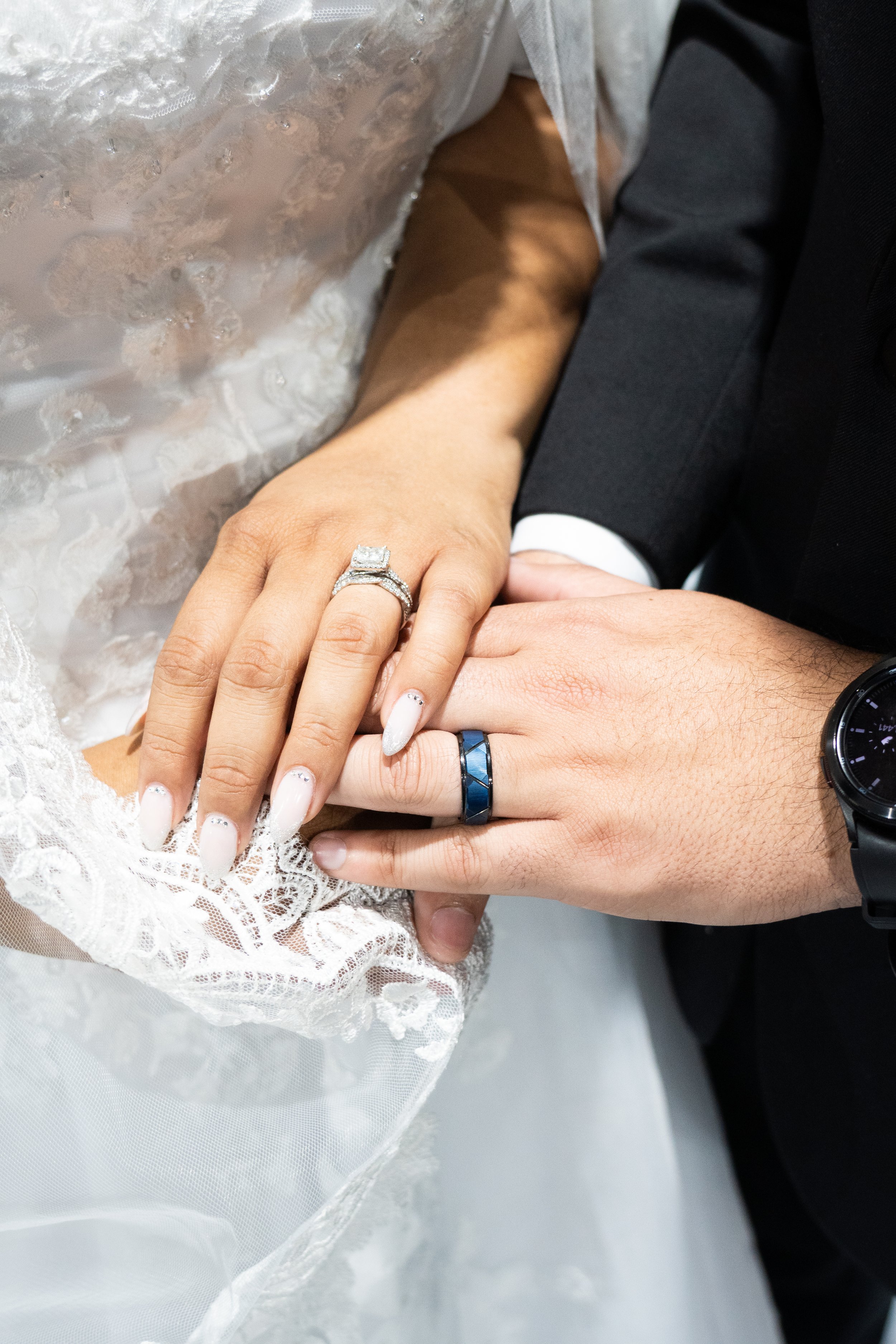 Close-up of a bride and groom holding hands, showing wedding rings, on their wedding day. The bride's hand is adorned with a diamond engagement ring and wedding band, and her nails are painted white with small embellishments. The groom's hand wears a