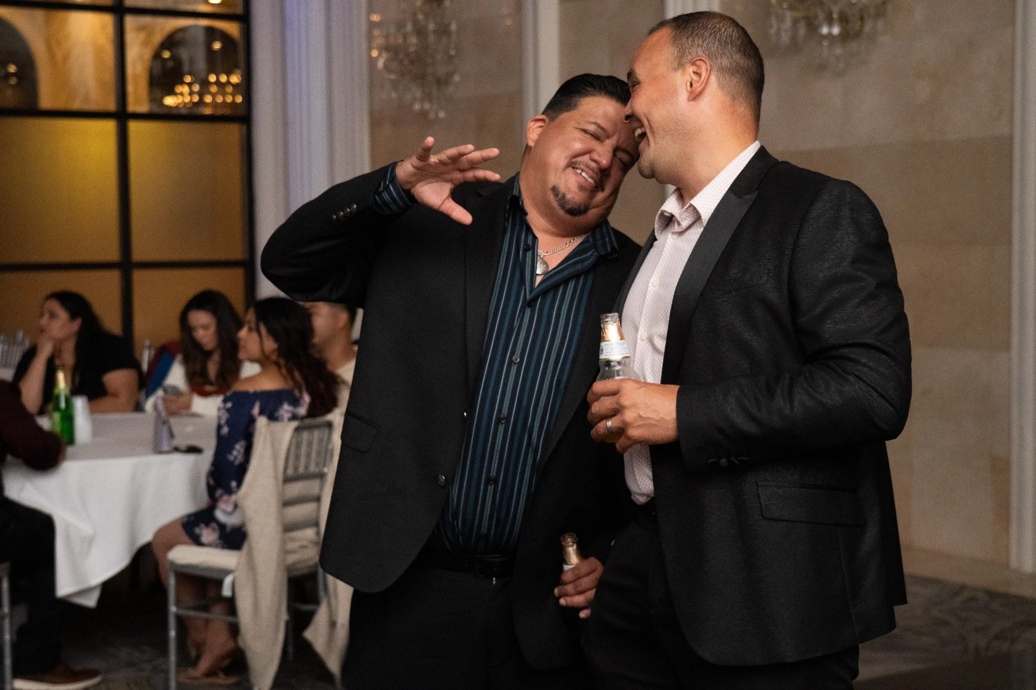 Two men in tuxedos smiling and touching foreheads at a social gathering, one holding a beer bottle, celebrating at a wedding reception.