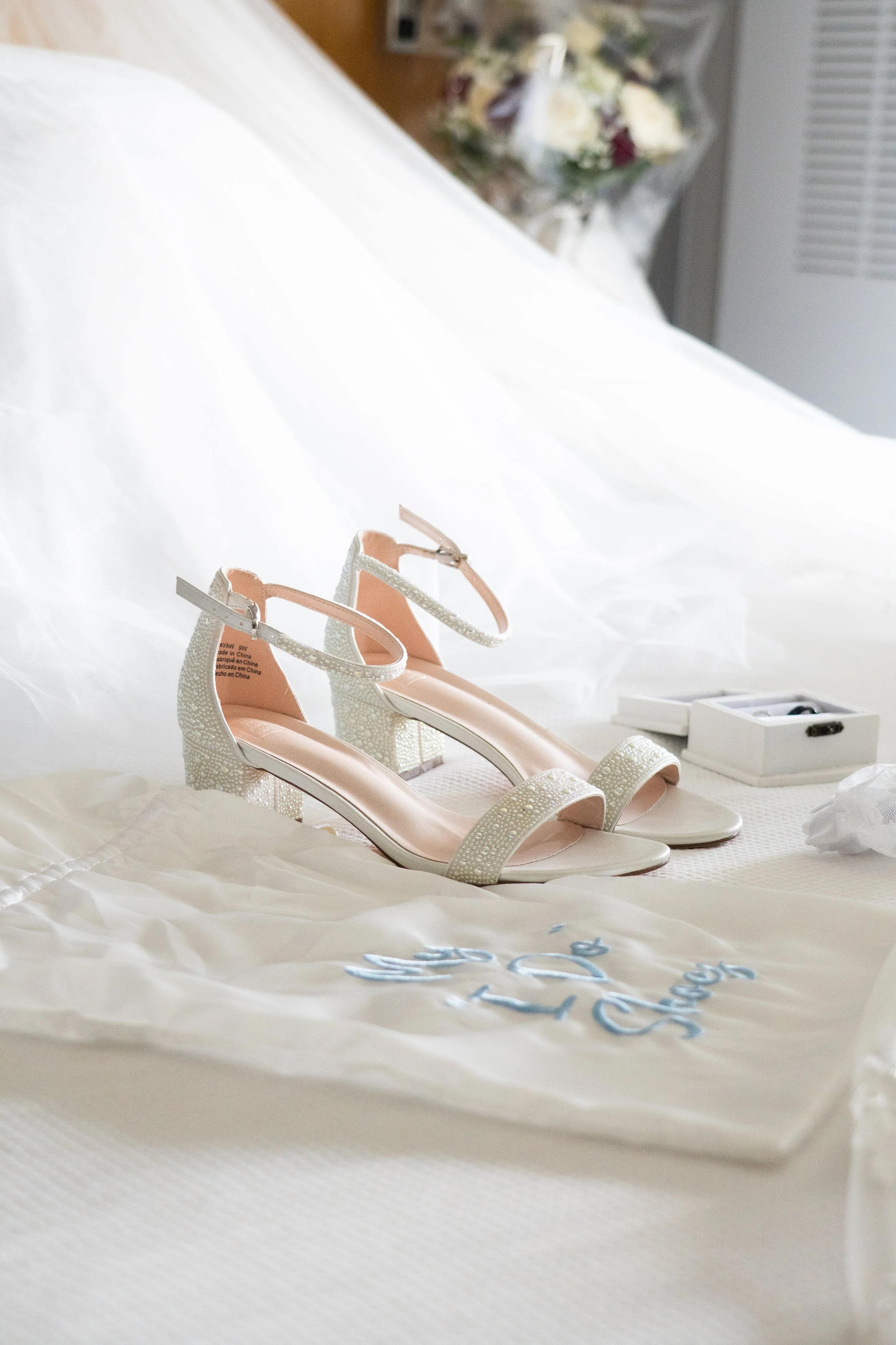 A pair of elegant white high-heeled shoes with ankle straps, decorated with small pearls or beads, placed on a white cloth with blue embroidery, on a bed with a white sheet and a bouquet of flowers in the background.