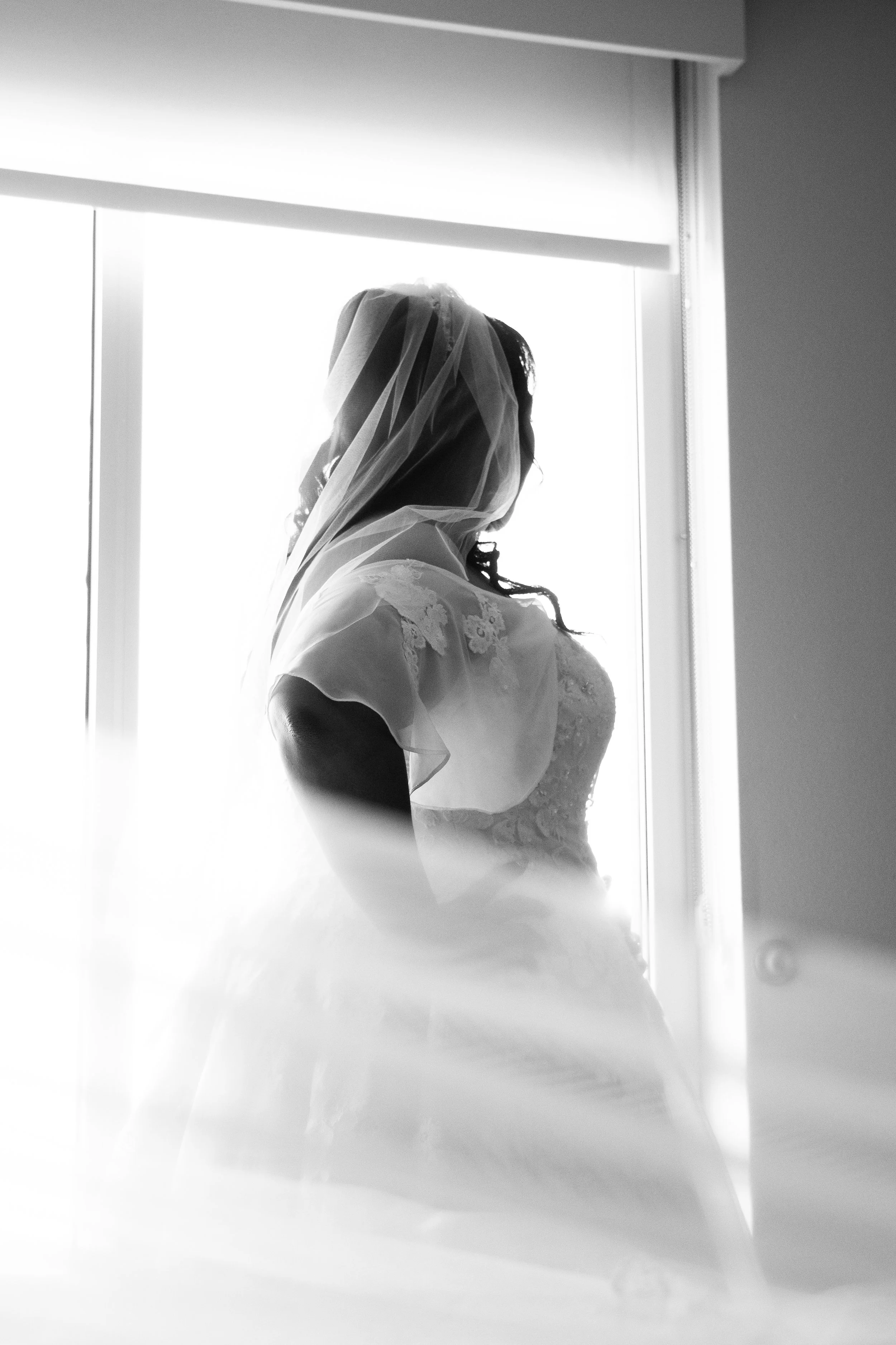 Silhouette of a bride in a wedding dress with a veil, standing near a bright window.