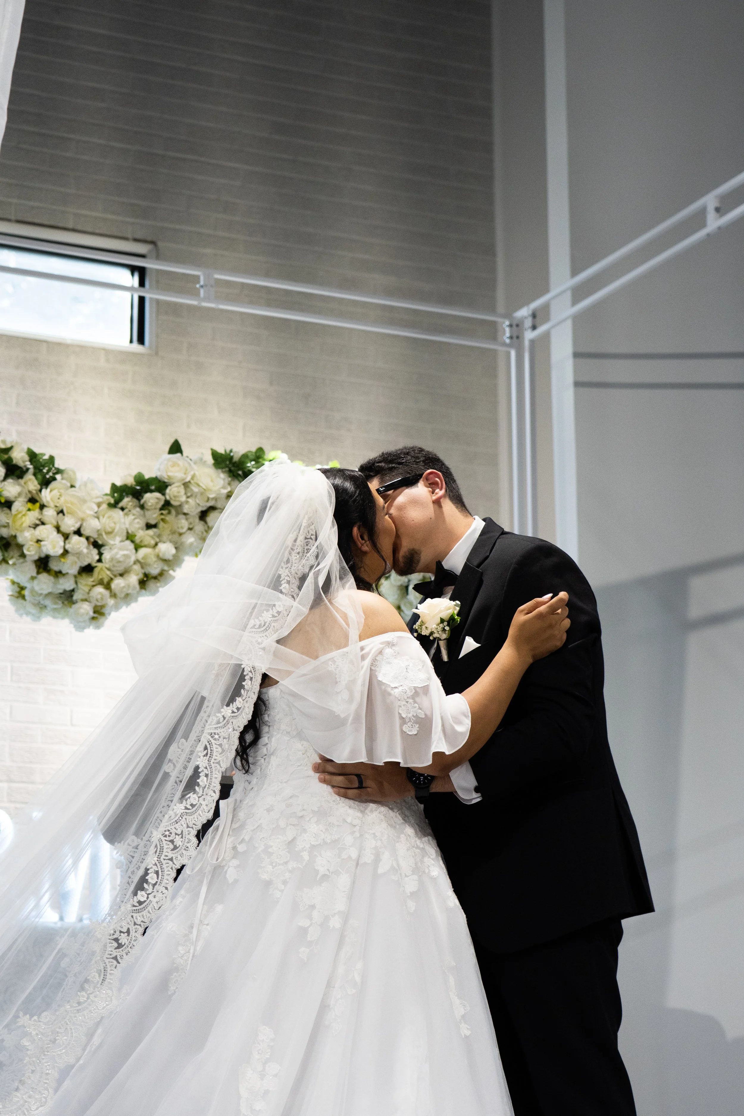 A bride and groom share a kiss during their wedding ceremony inside a modern venue with a floral backdrop.