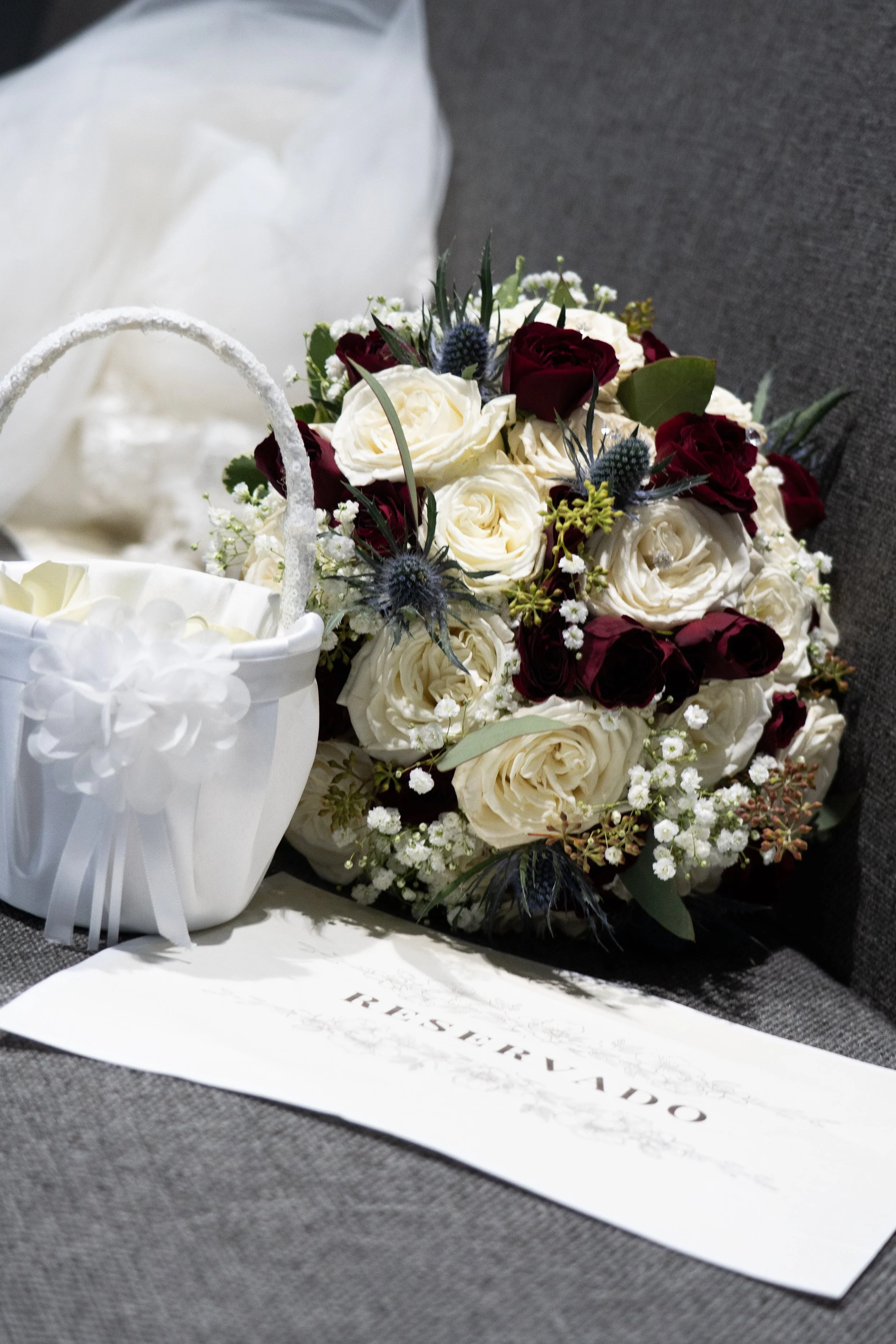 A wedding bouquet of white and dark red roses, mixed with greenery and small white filler flowers, placed next to a white basket decorated with a white flower and ribbon, on a dark fabric surface.
