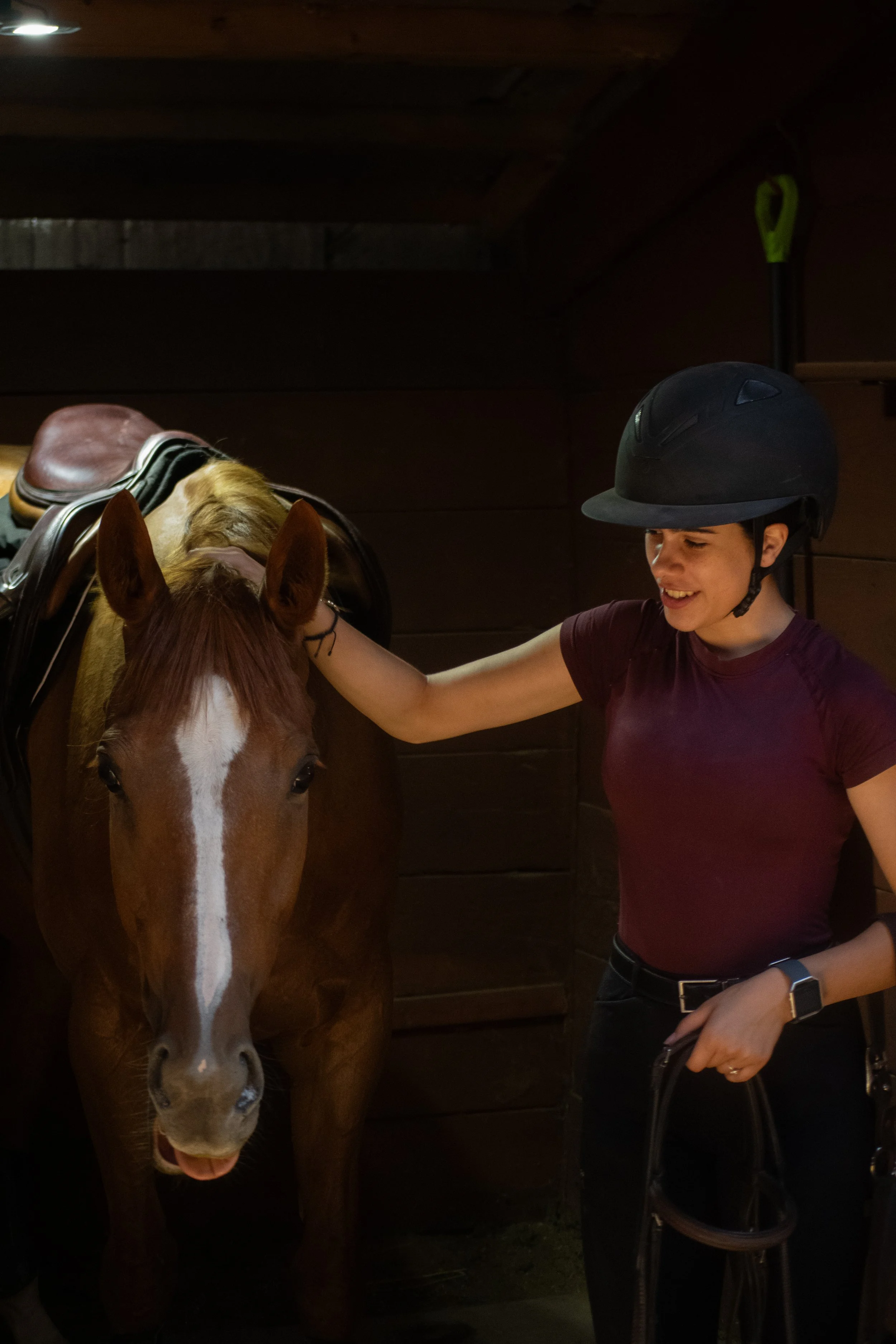A woman wearing a riding helmet smiles as she stands next to a brown and white horse with a saddle, inside a wooden stable.