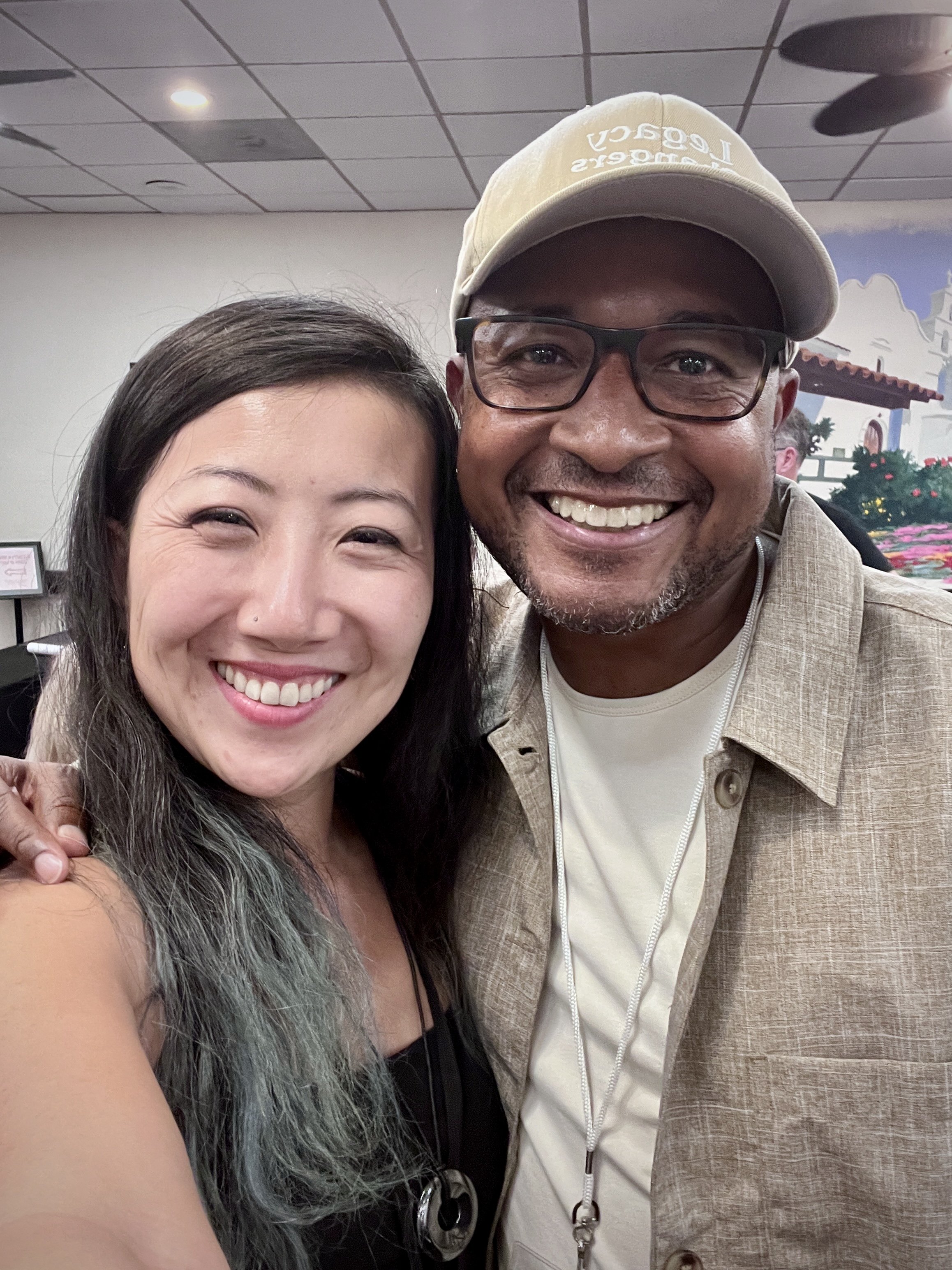 A woman and a man smiling and posing together for a selfie indoors. The woman has straight dark hair with light-colored tips and the man wears glasses and a beige hat.