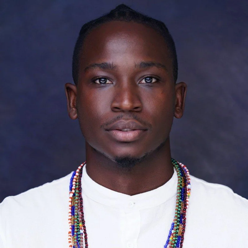 Portrait of a young man with dark skin, short hair, wearing a white shirt and multiple colorful beaded necklaces, against a dark background.