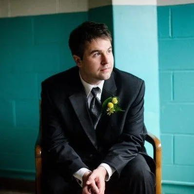 A young man in a suit sitting on a chair against a teal wall.