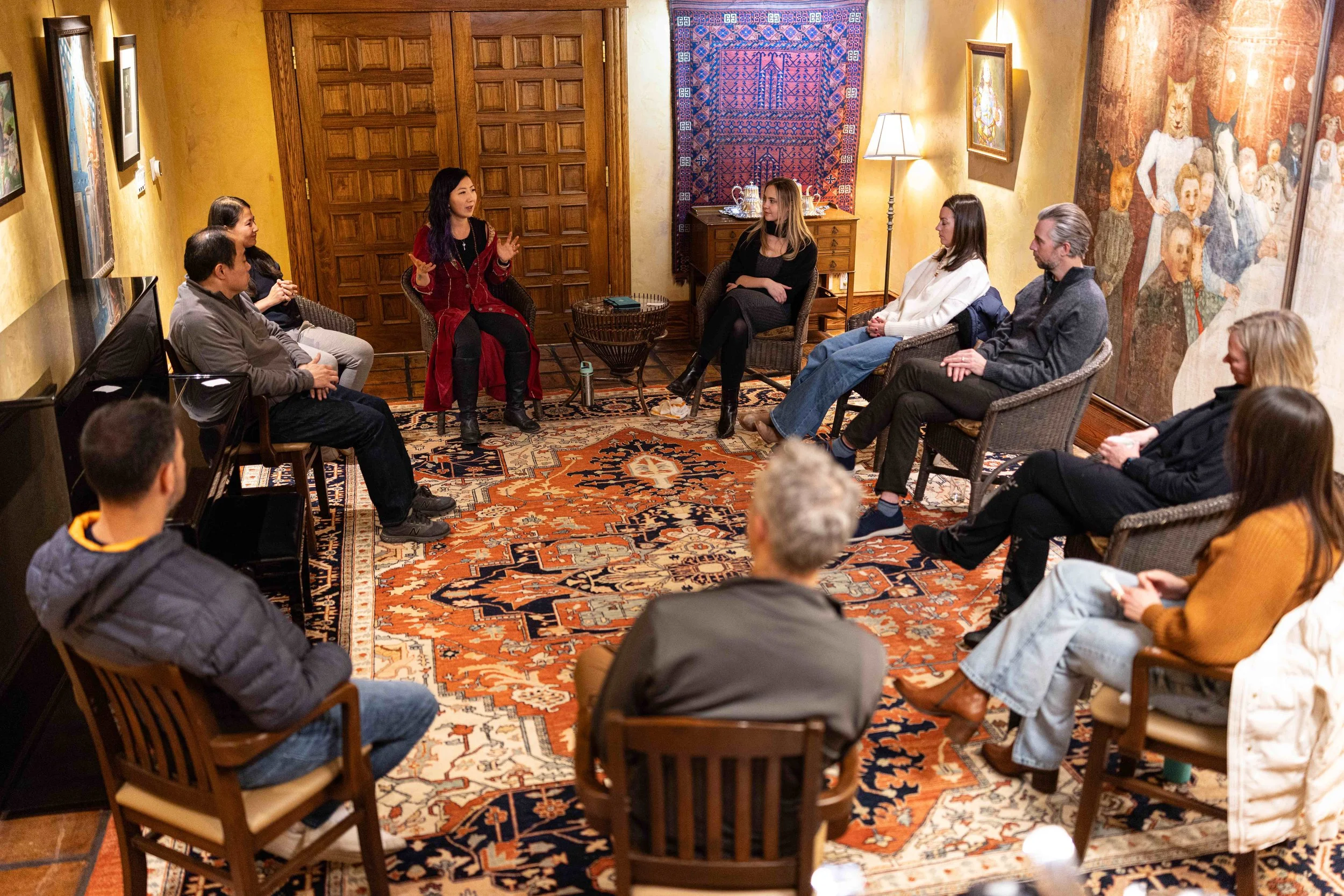 A group of people sitting in a circle in an art gallery or museum, engaging in a discussion or workshop, with artworks on the walls and a patterned rug on the floor.