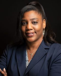 Professional portrait of a woman with dark hair in a ponytail, wearing a navy blazer, pearl necklace, and earrings, against a black background.