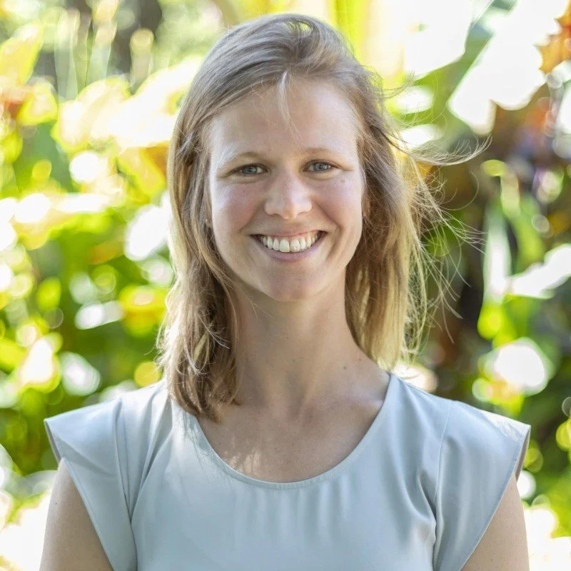 A smiling woman with shoulder-length light brown hair standing outdoors in front of green leafy plants.