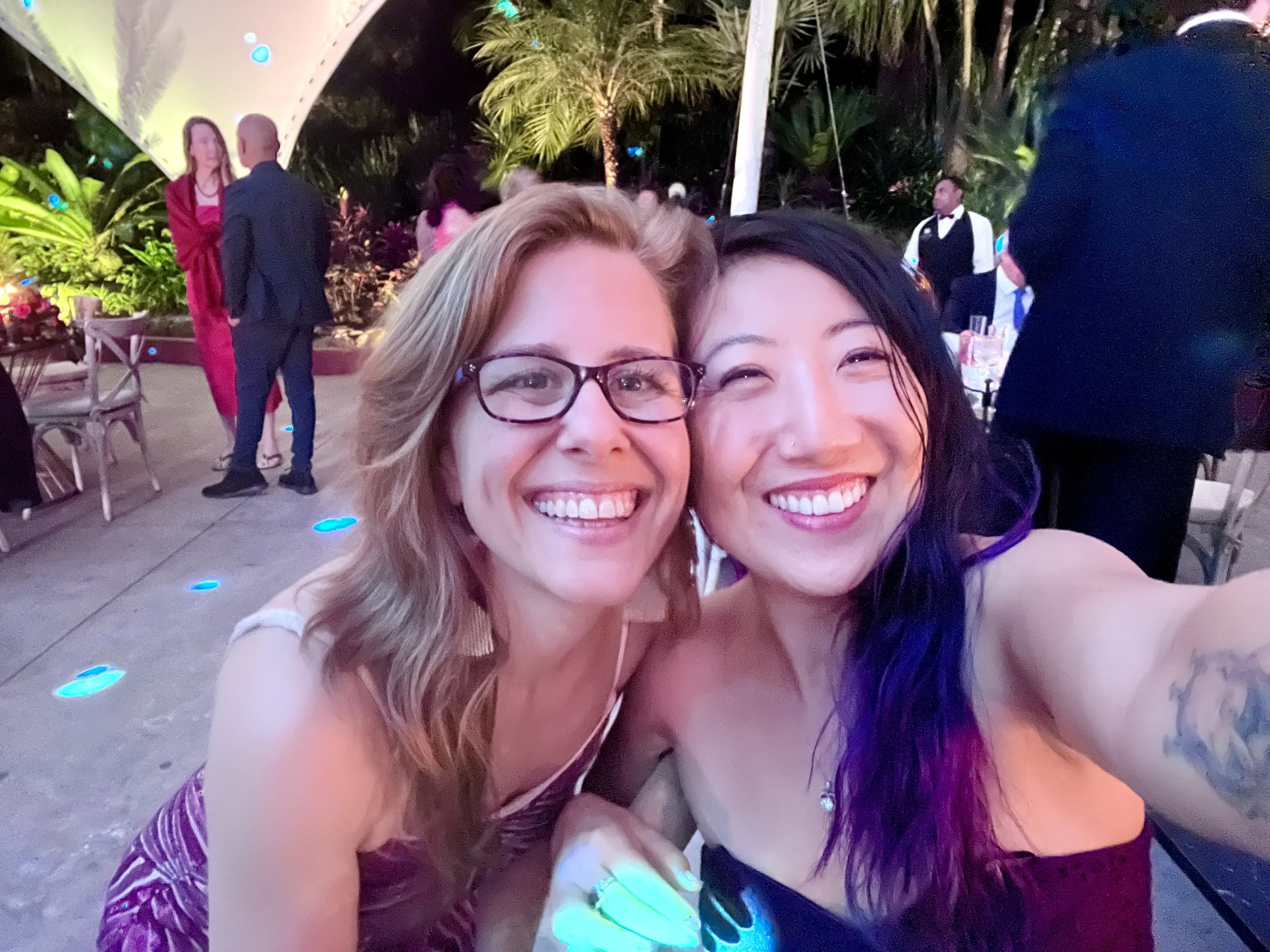 Two women smiling and taking a selfie at an outdoor evening event, with other guests and lush tropical plants in the background.