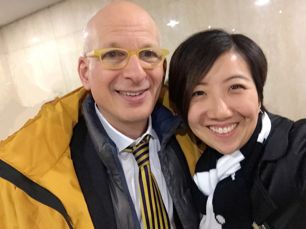 A man and a woman taking a selfie together, both smiling, indoor setting with beige walls.