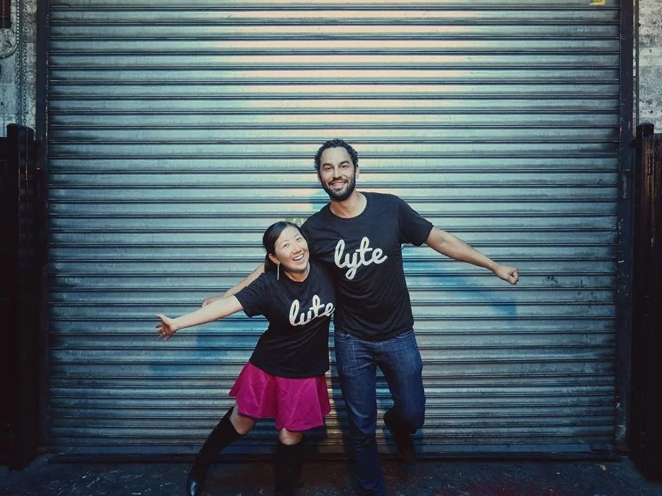 Two people smiling and posing in front of a metal roll-up door, both wearing black t-shirts with the word "lyfe" on them. The woman is wearing a pink skirt and black knee-high socks, and the man is wearing blue jeans.