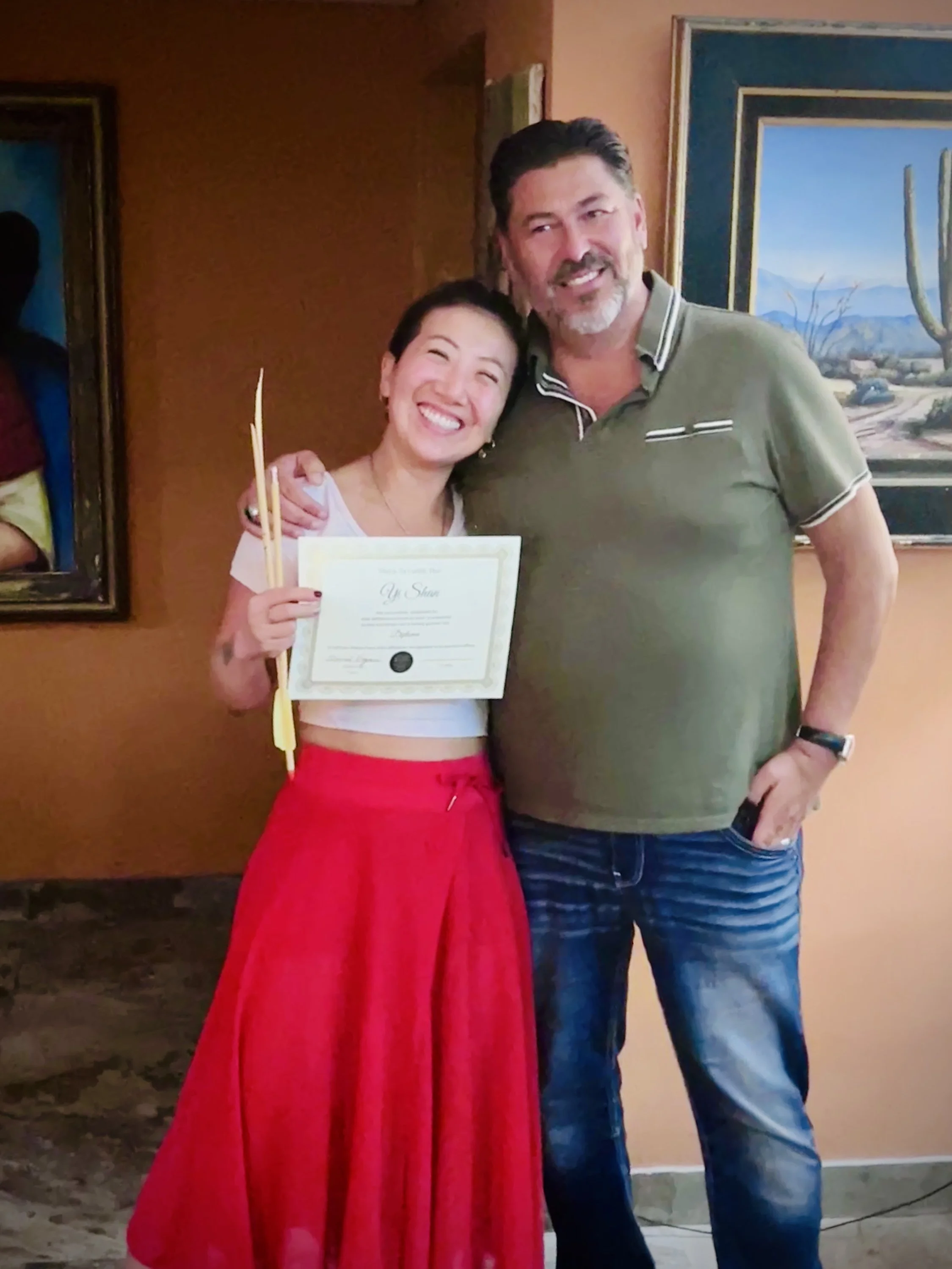A woman holding a diploma and chopsticks smiling with a man with gray hair in a green polo shirt, standing in front of desert landscape paintings on a brown wall.