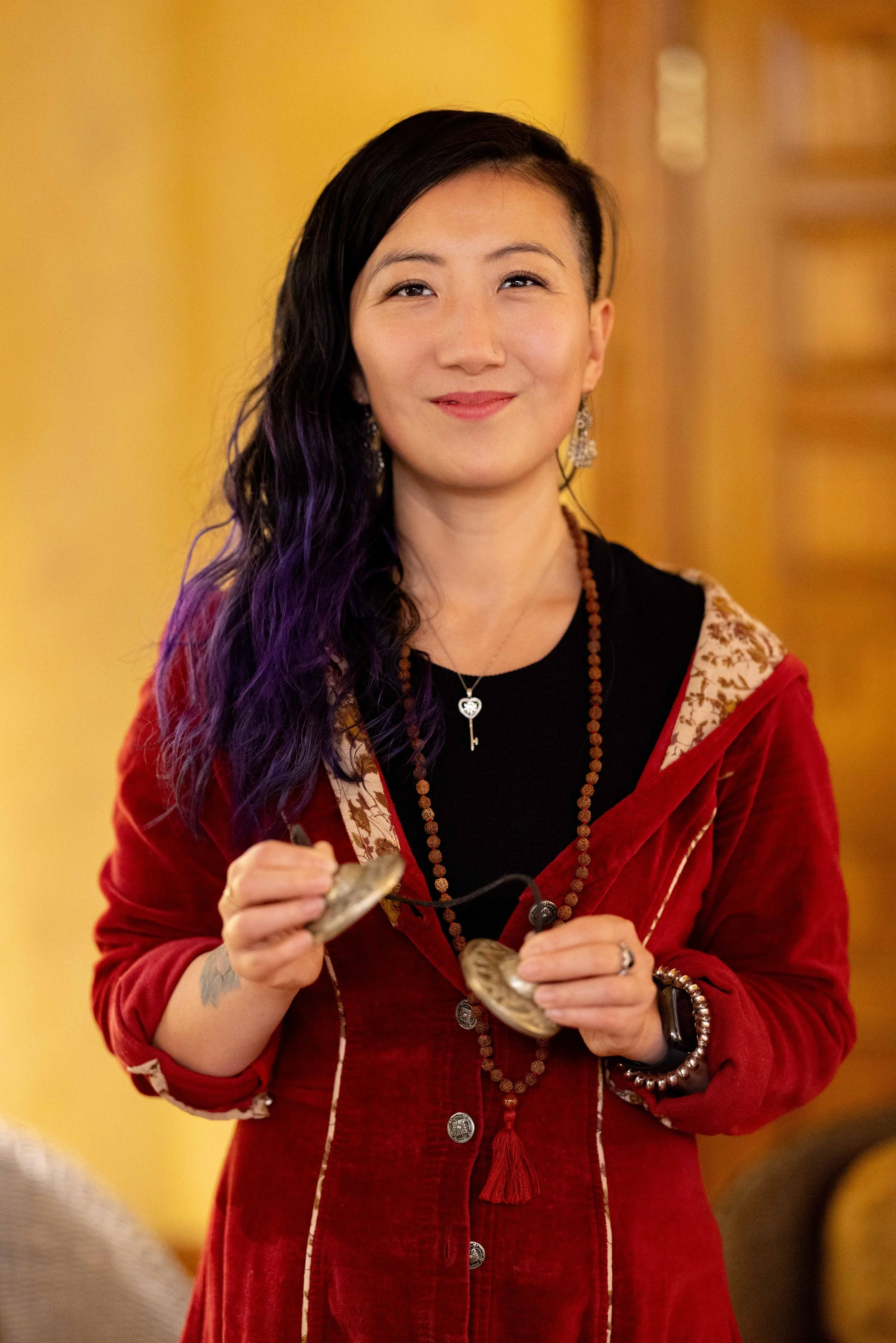 A woman with black and purple wavy hair wearing a red velvet jacket with embroidered details, holding a pocket watch, and smiling at the camera.