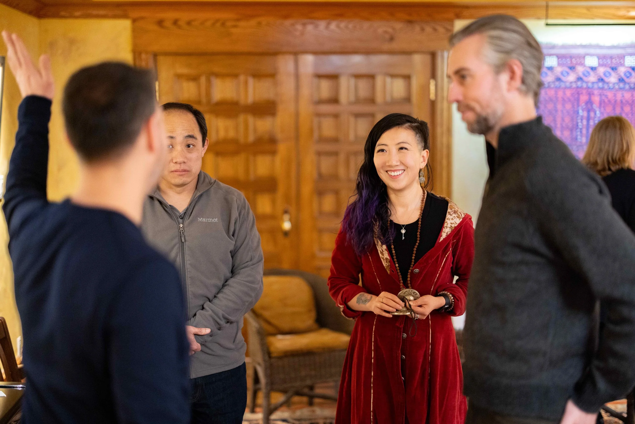 Four people engaged in conversation in a warmly decorated room. One woman, smiling, wears a red velvet jacket and holds objects, while another man is turned away, and two other men look on.