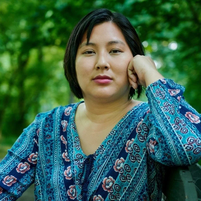 A woman with short black hair wearing a blue patterned top sitting outdoors surrounded by green foliage.