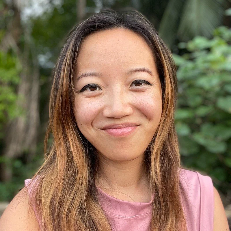 A woman with long, wavy hair smiling outdoors with green foliage in the background.