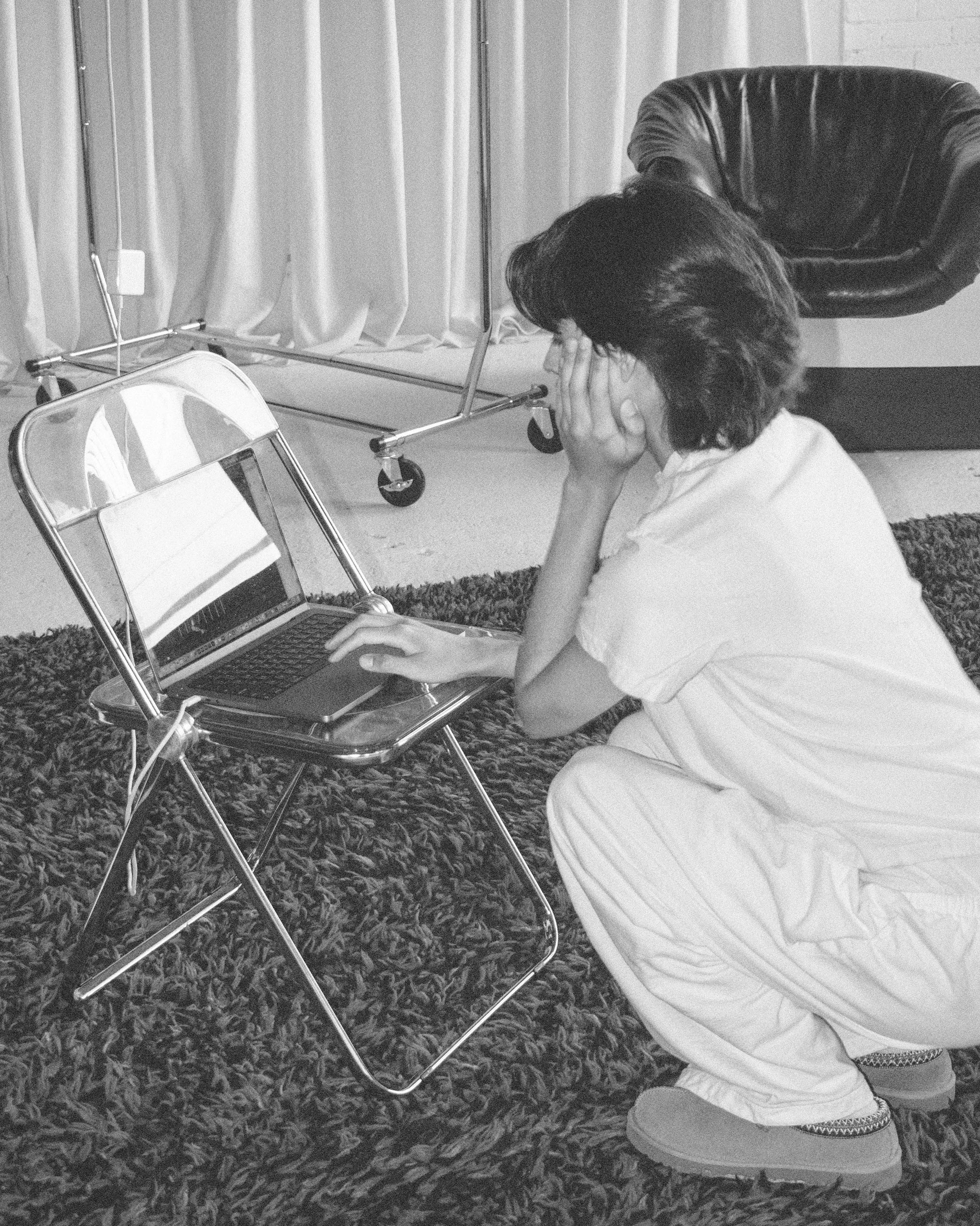 Person sitting on a carpeted floor, looking at a laptop on a small metal table, with hands on their face, appearing distressed or upset.