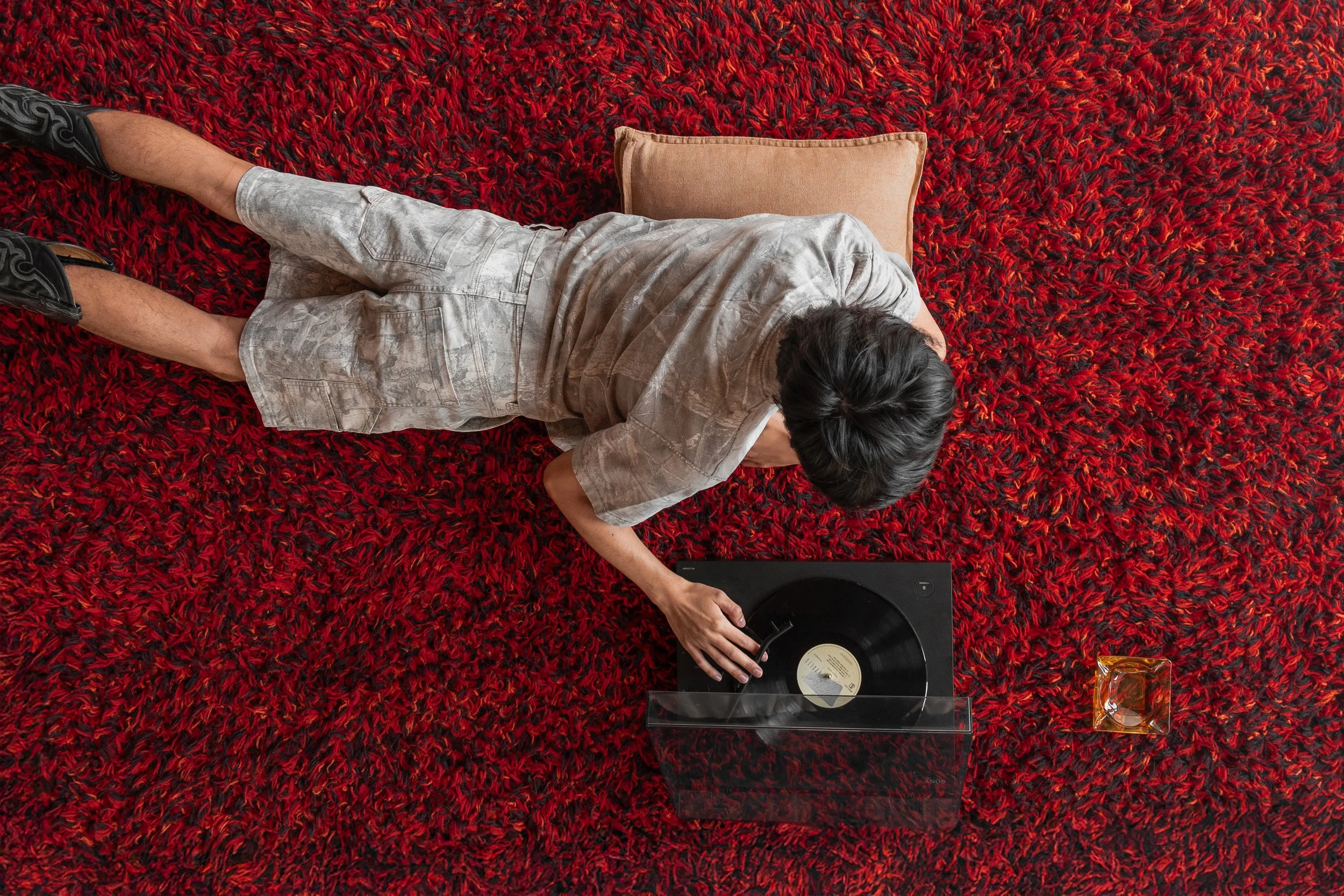 Person lying on red shag carpet, listening to vinyl record on turntable, with a pillow and a glass ash tray nearby.