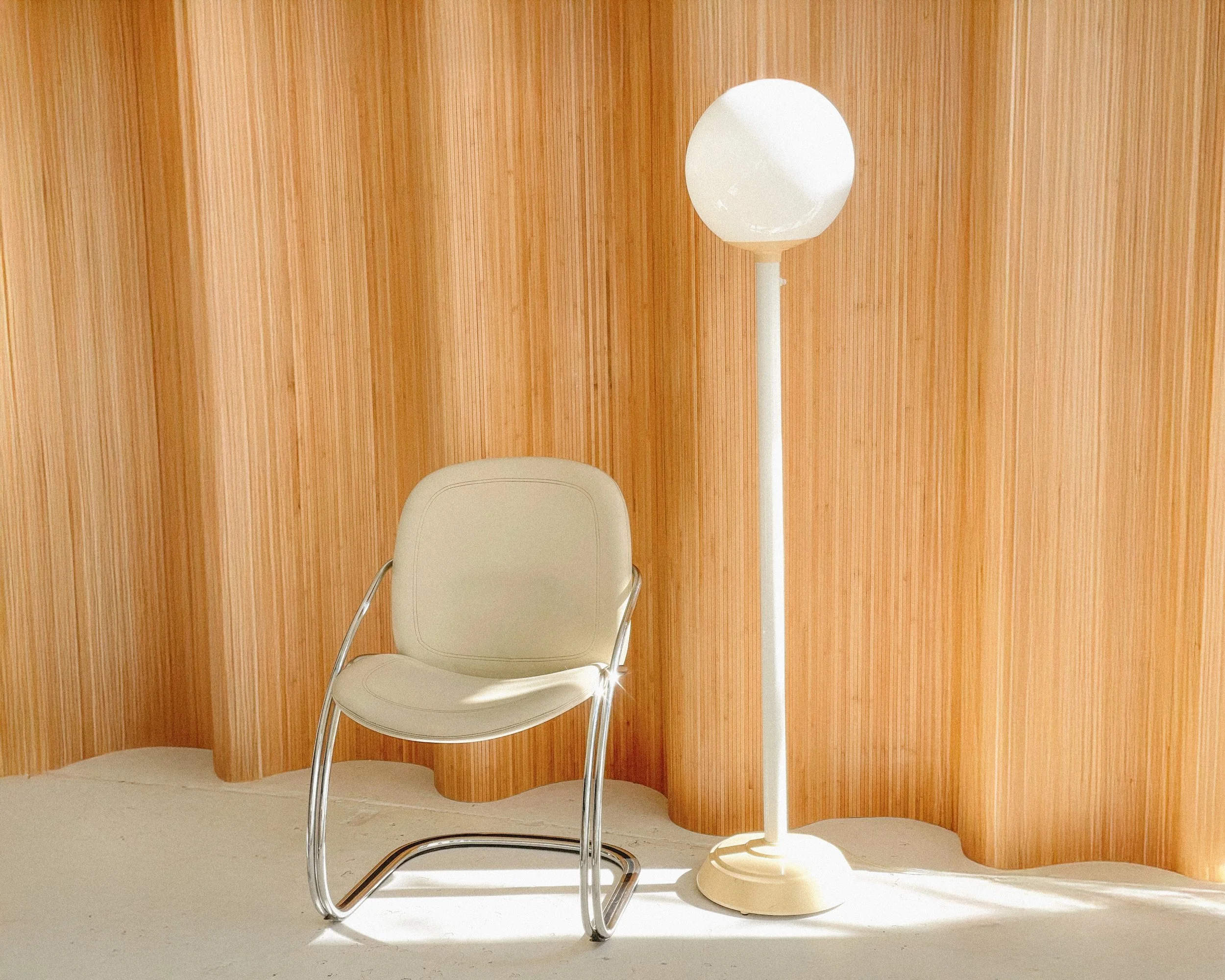 A white chair with metal armrests and legs beside a tall white floor lamp with a round shade, in front of a wooden-paneled wall.