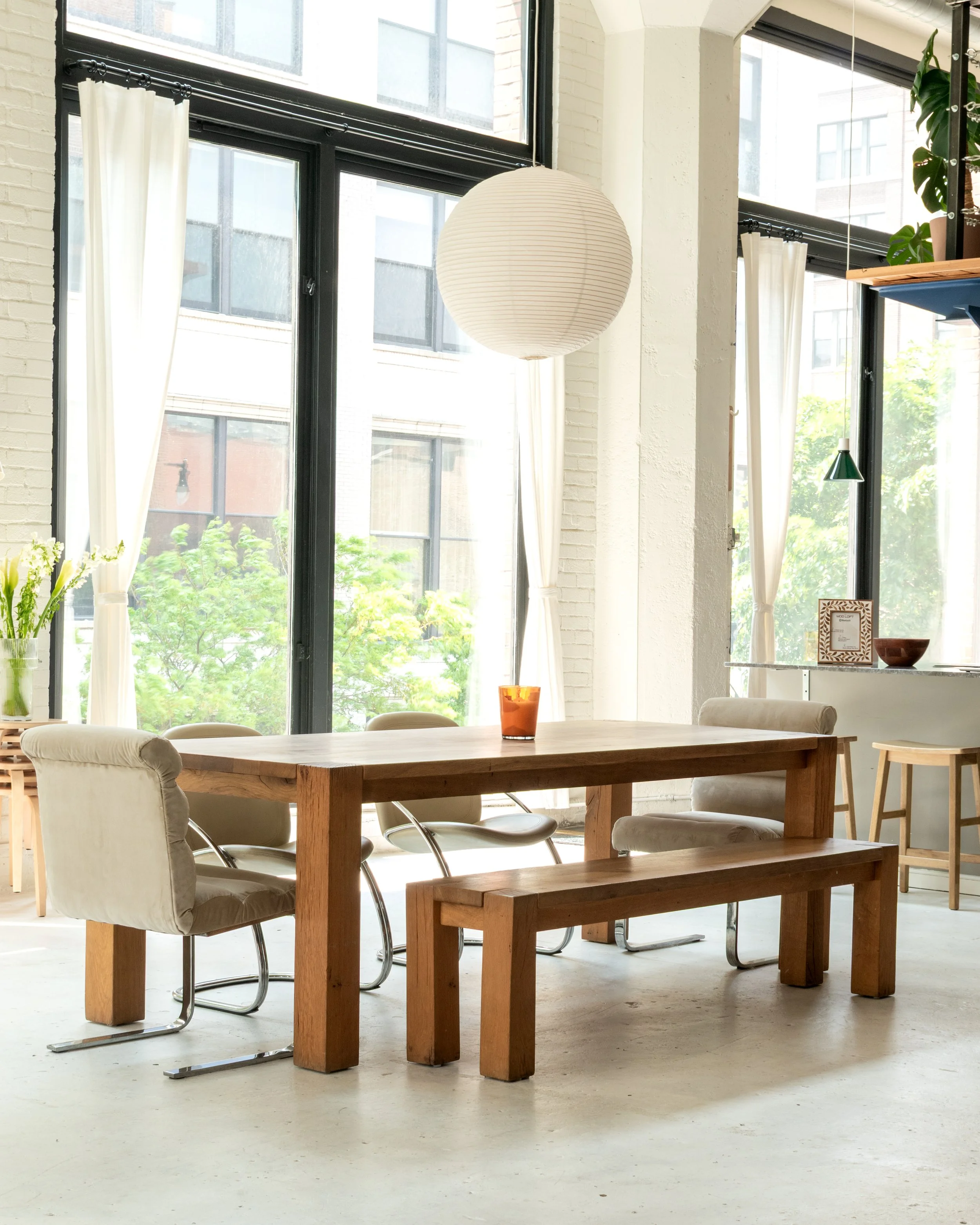 A bright, modern dining area with a wooden table, cream cushioned chairs, a wooden bench, and large windows with curtains letting in natural light.