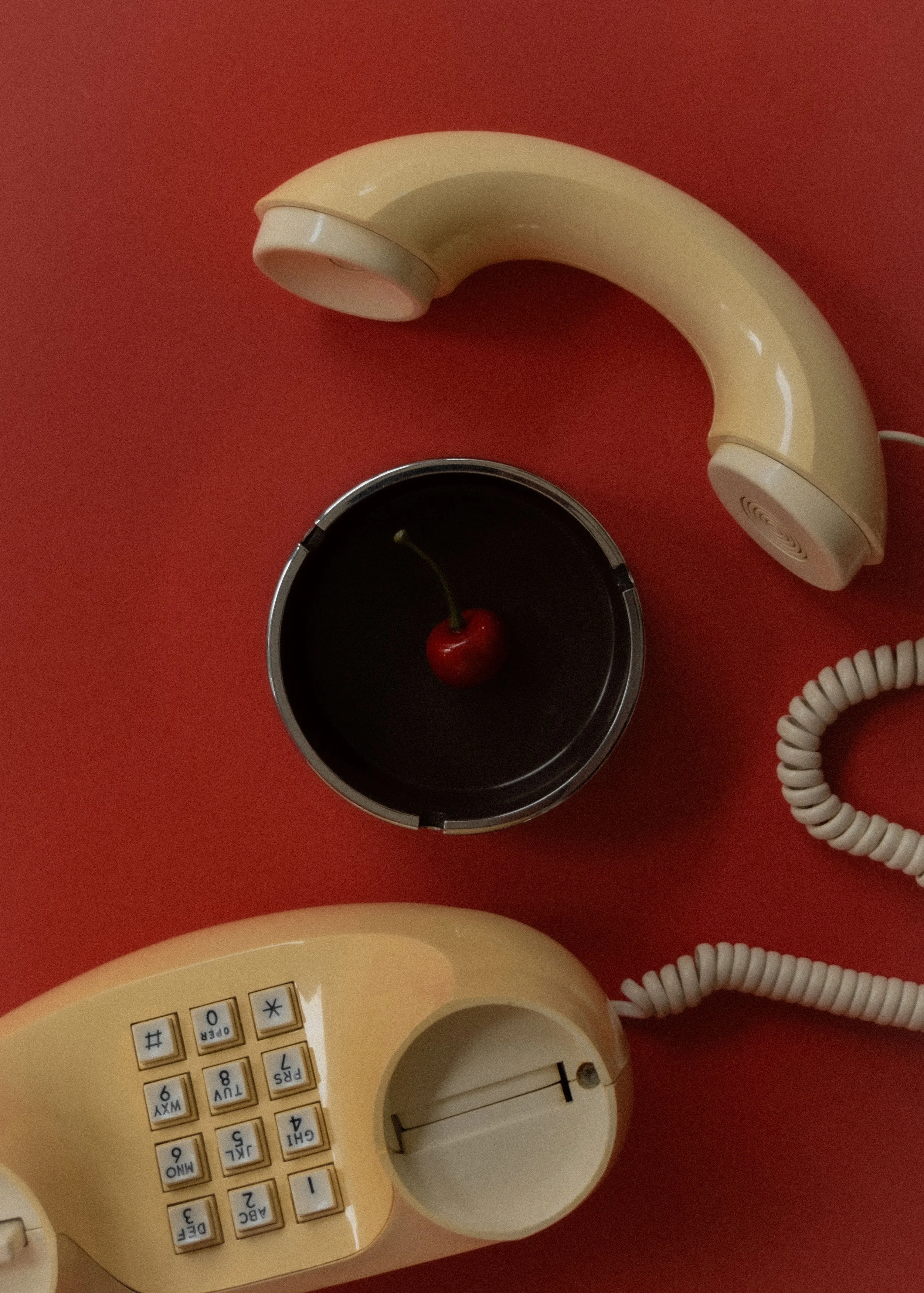 A vintage beige rotary telephone, a beige cordless phone, and a small black dish with a cherry on top are arranged on a red surface.