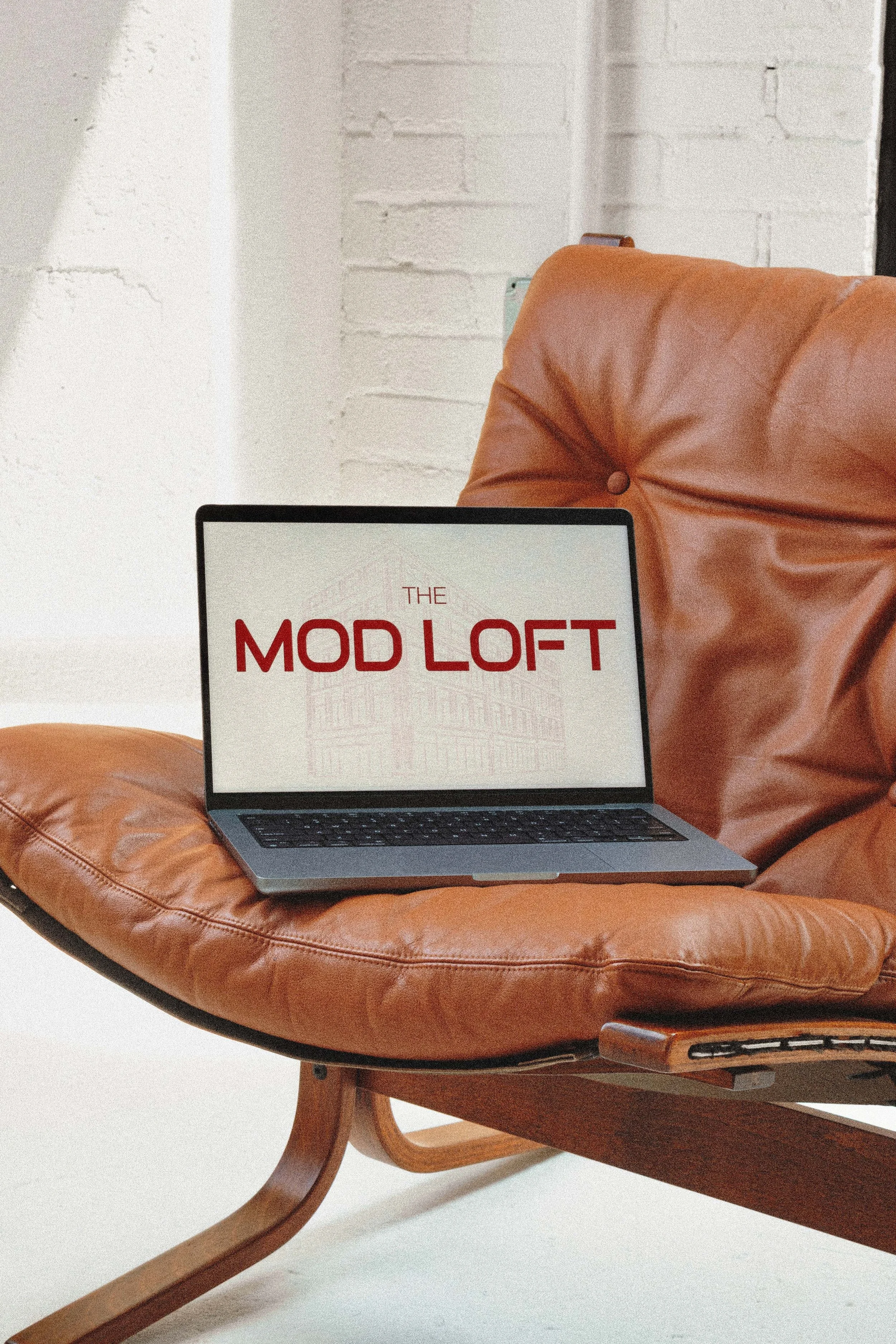 Laptop on a brown leather Siesta Lounge chair displaying the text 'The MOD LOFT' with a faint building outline in the background.