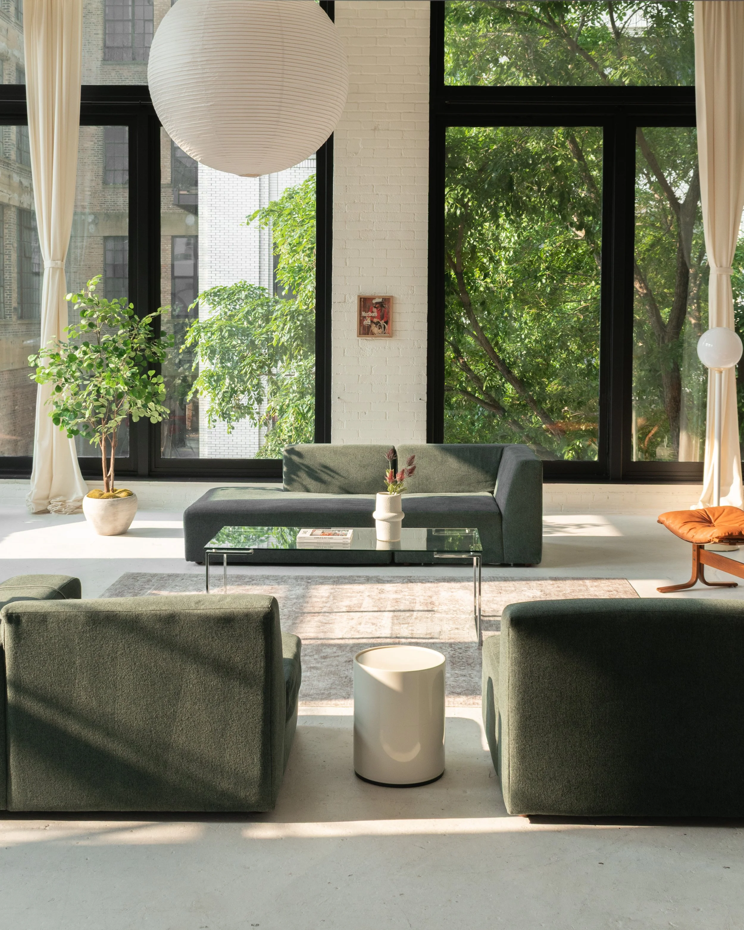 Modern living room with large windows, white brick wall, green sofa, armchairs, coffee table, potted plants, and natural light.