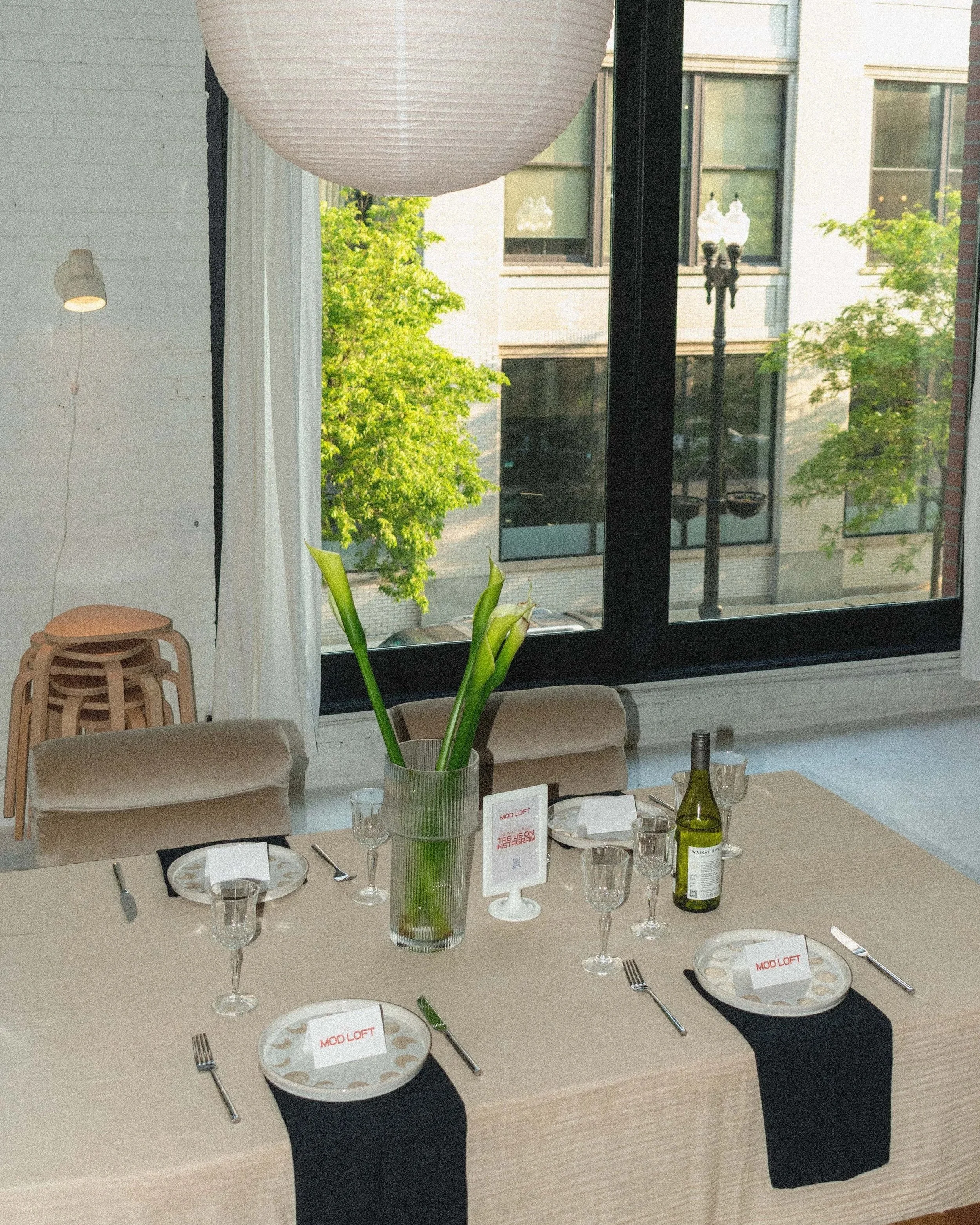 A dining table set near a large window with greenery outside. The table has plates, glasses, silverware, and a vase with green plants, possibly calla lilies. There are small chairs at the table, and stacked wooden stools in the background.