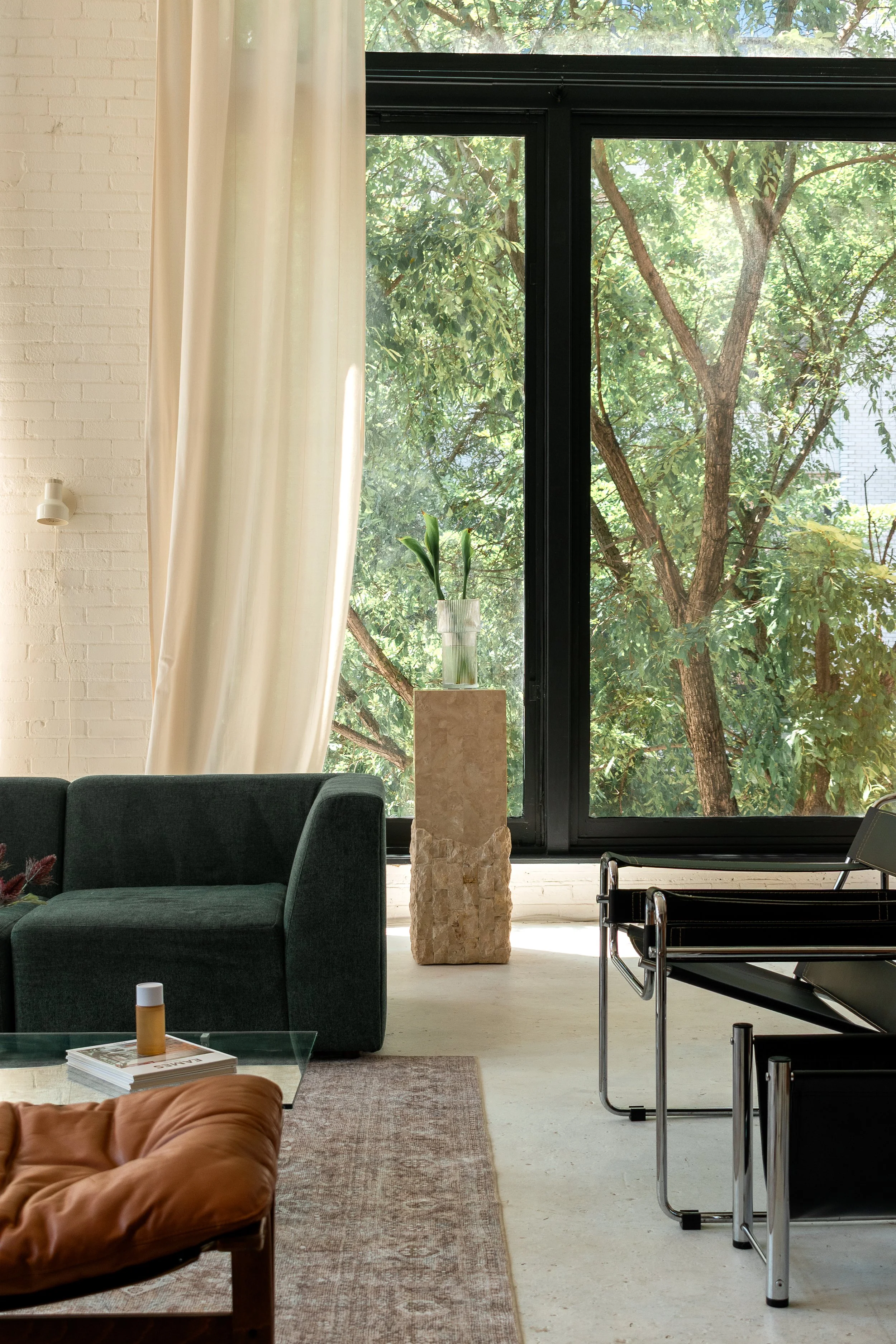 Living room with large window showing leafy trees outside, dark green sofa, stack of magazines, orange cushion, glass coffee table, decorative vase with green plants on stone pedestal, cream curtain, white brick wall.