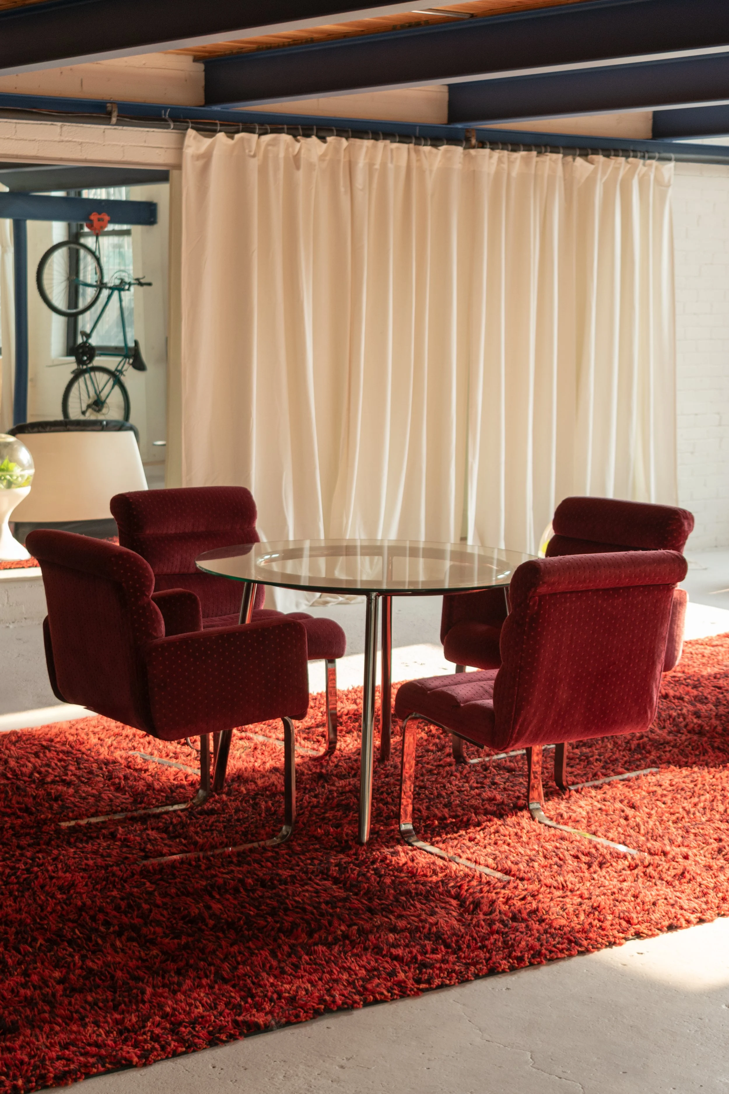 A vintage round glass table surrounded by four red upholstered chairs on a red shaggy rug, with white curtains in the background, in a room with white brick walls and a bicycle hanging on the wall.