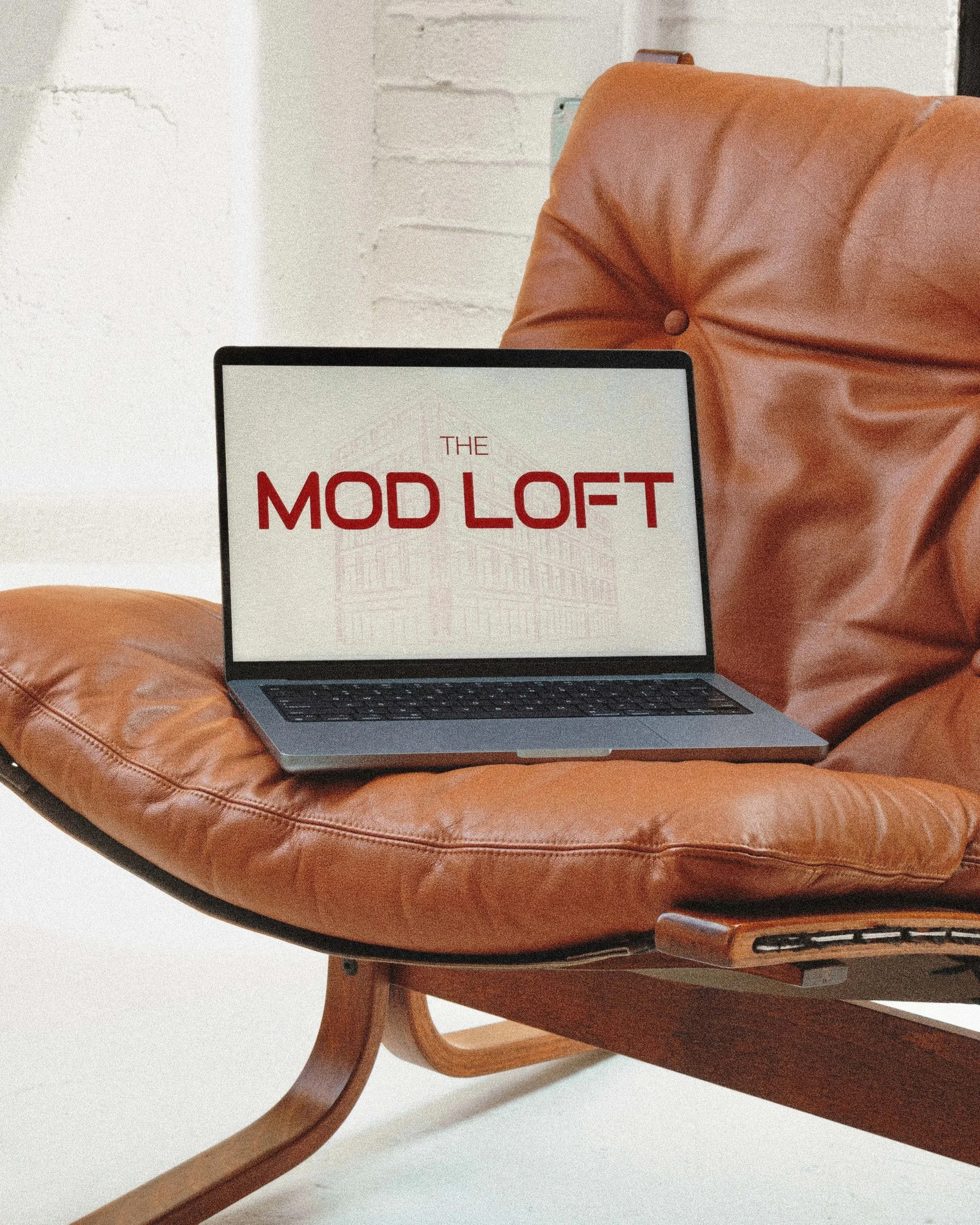 Laptop on a brown leather chair displaying the text 'THE MOD LOFT' in red.