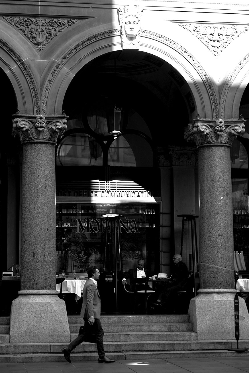 Black-and-white photo of a building with large arched windows and ornate columns. Inside, two men are seated at a table, and a man in a suit is walking past the steps in front of the building.