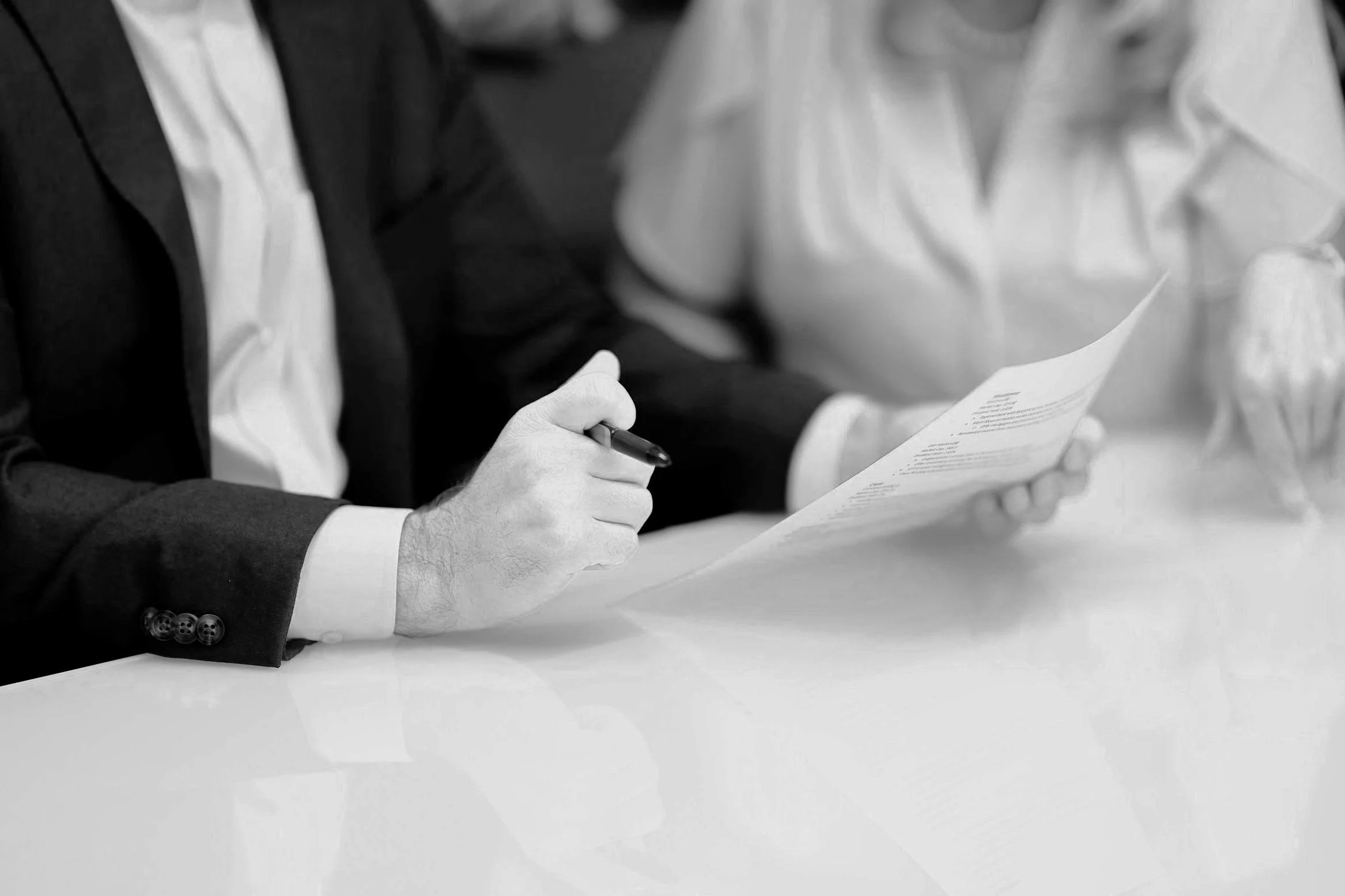 A man in a suit and white shirt sitting at a table, holding a pen and reading a document, with a woman in a white blouse sitting nearby.