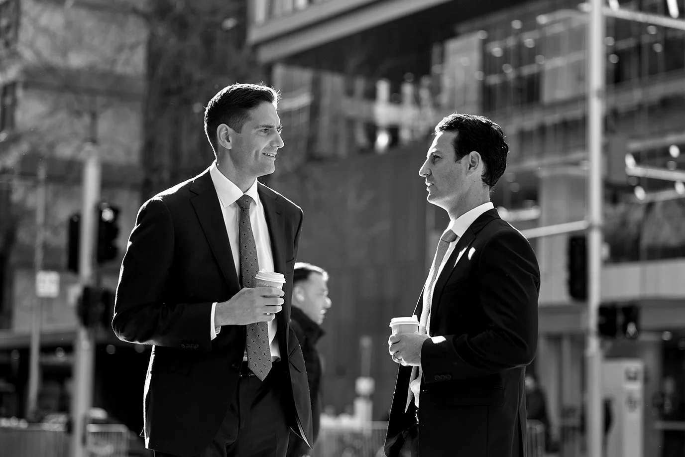 Two men in suits having a conversation outdoors, each holding a coffee cup, in a city setting with modern buildings in the background.