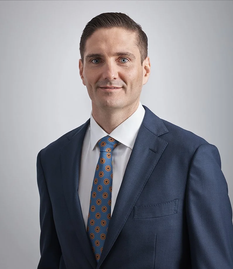 Professional portrait of a man in a navy suit with a white shirt and a colorful patterned tie, standing against a plain light gray background.