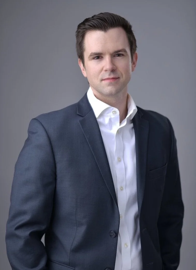 A young man in a dark suit and white shirt posing against a gray background.