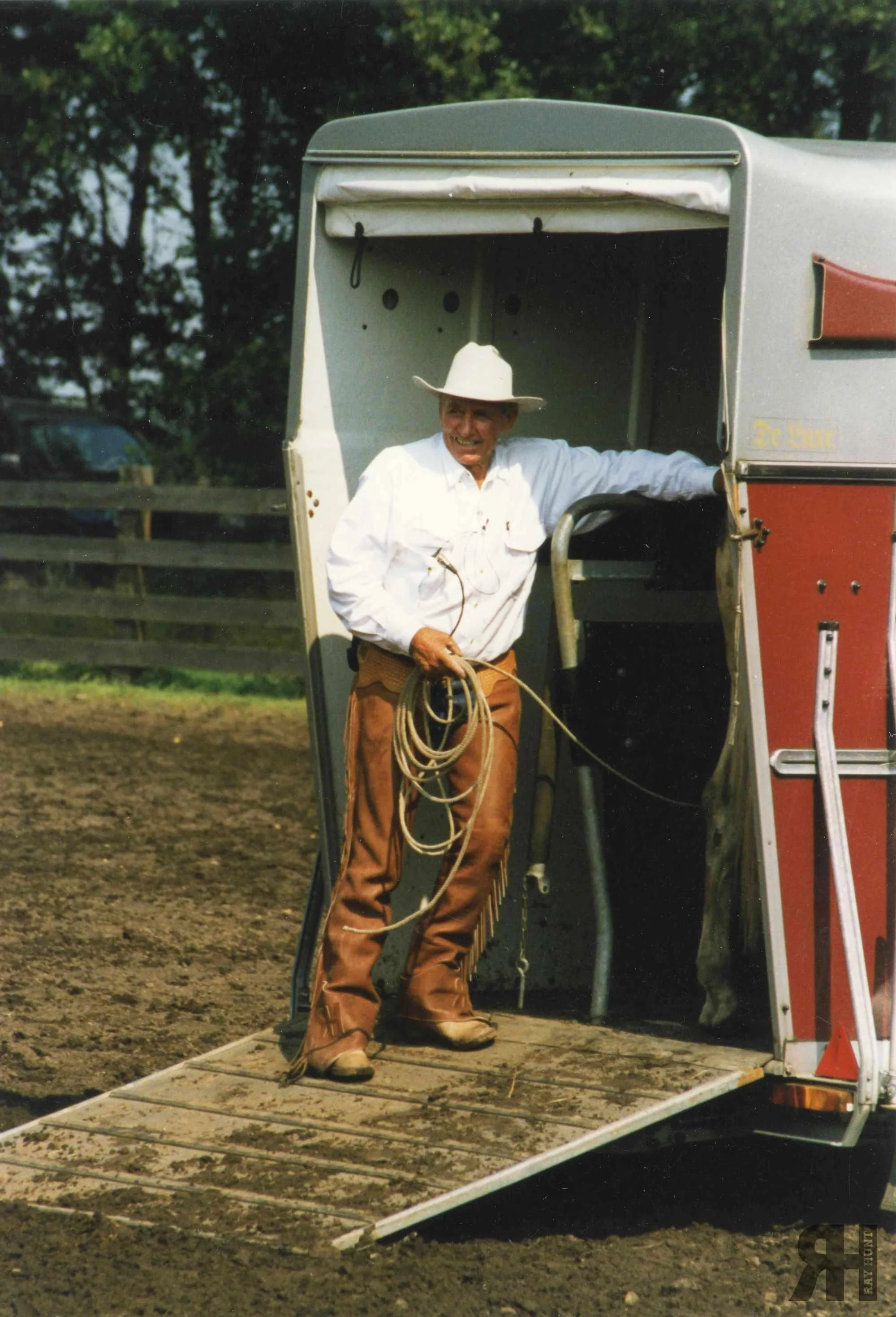 Ray Hunt on the trailer ramp with a succesfully loaded horse during a trailer loading demo. He is smiling, wearing a crisp white shirt, leather chaps, signature white hat with rope in hand.