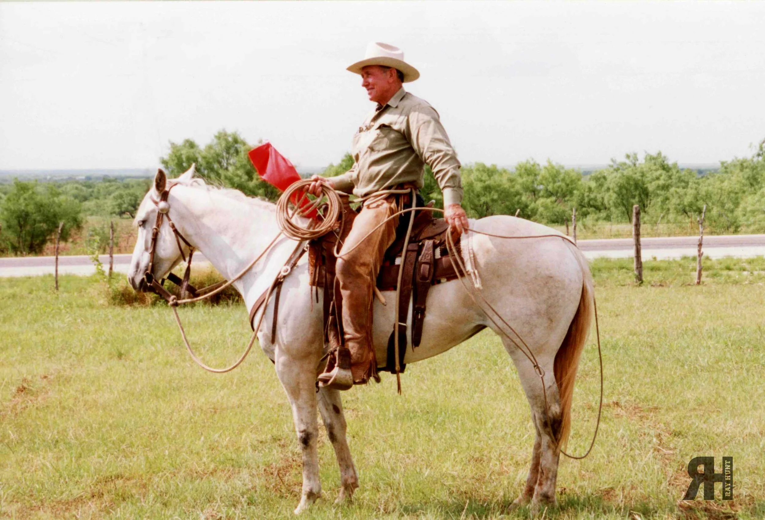 Ray Hunt, horseman, sitting atop a light colored horse with his signature orange flag in hand, lasso resting on the horse's rump, while the horse is so relaxed, she' dozing off in the sunshine. Photograph from the Ray and Caroly Hunt™ Family Archive.