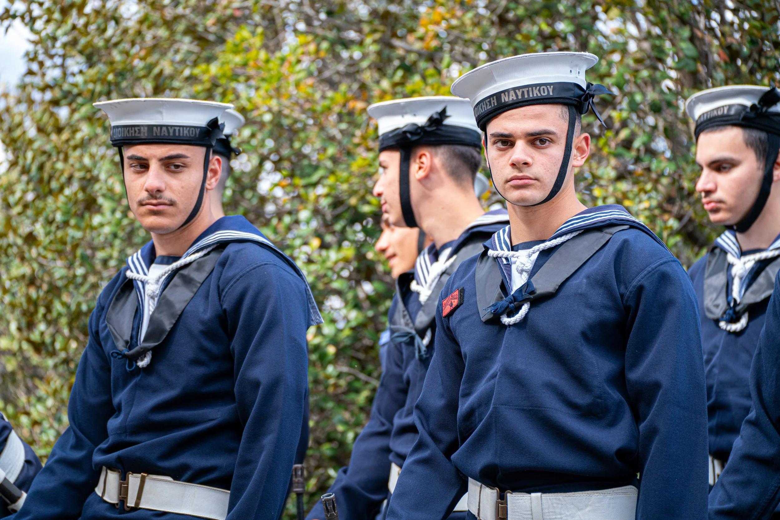 Group of young sailors in uniform standing outdoors, with greenery in the background.
