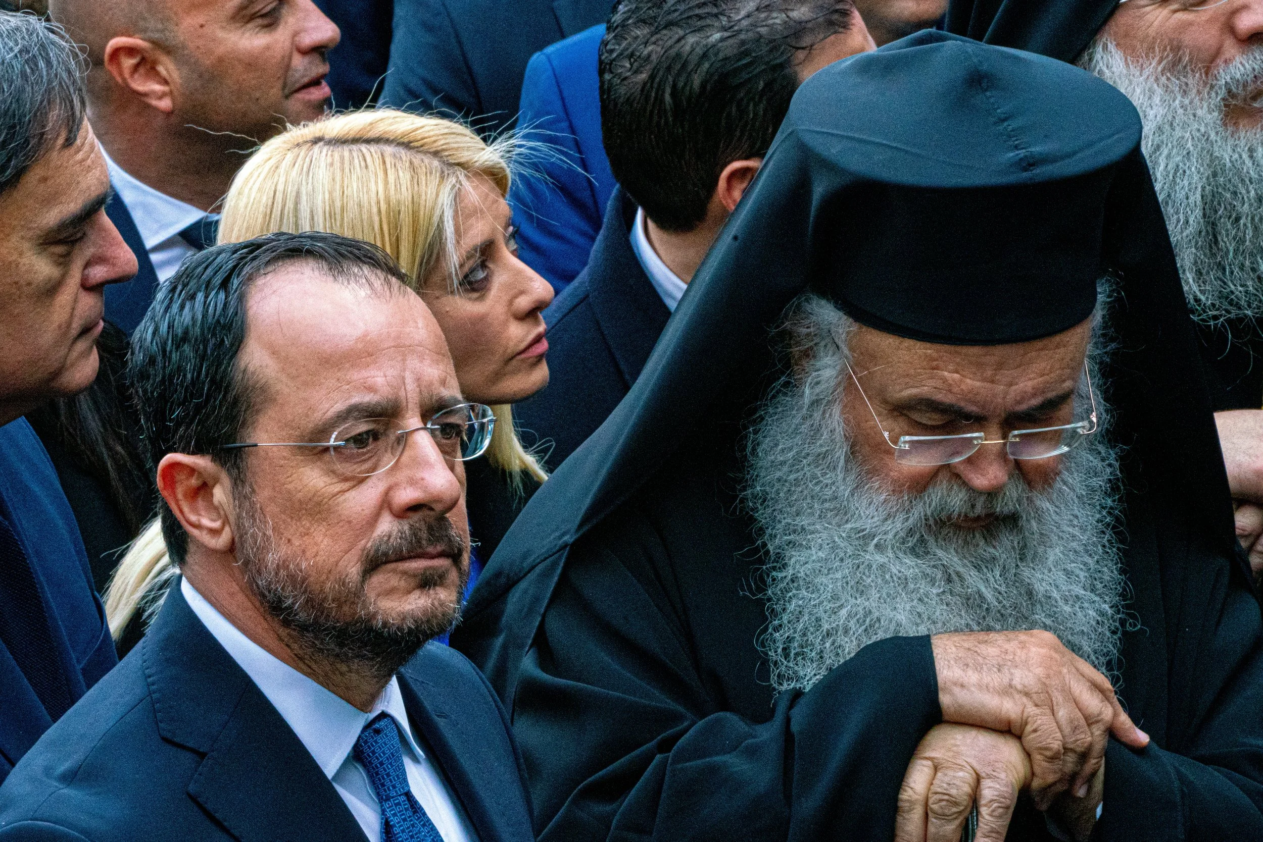 A group of people, including religious figures and individuals in suits, gathered in a solemn moment, with one man dressed as a religious bishop or priest with a large black head covering and long white beard, in an outdoor setting.