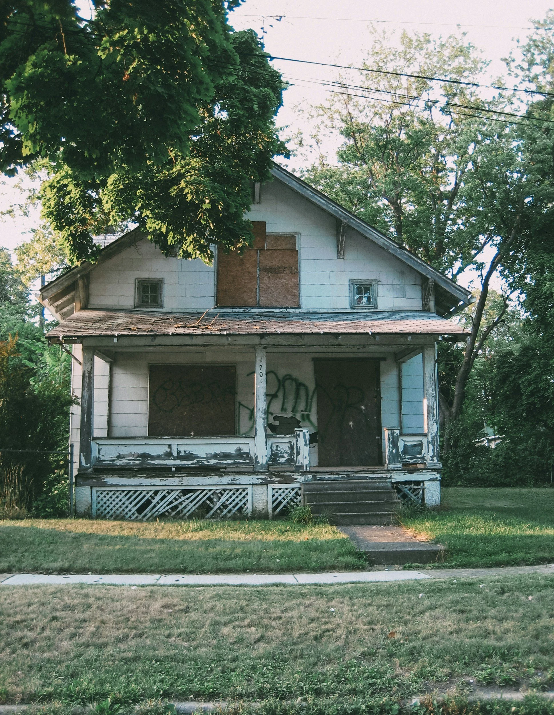 An abandoned house with boarded-up windows and graffiti on the front, surrounded by green grass, trees, and power lines.
