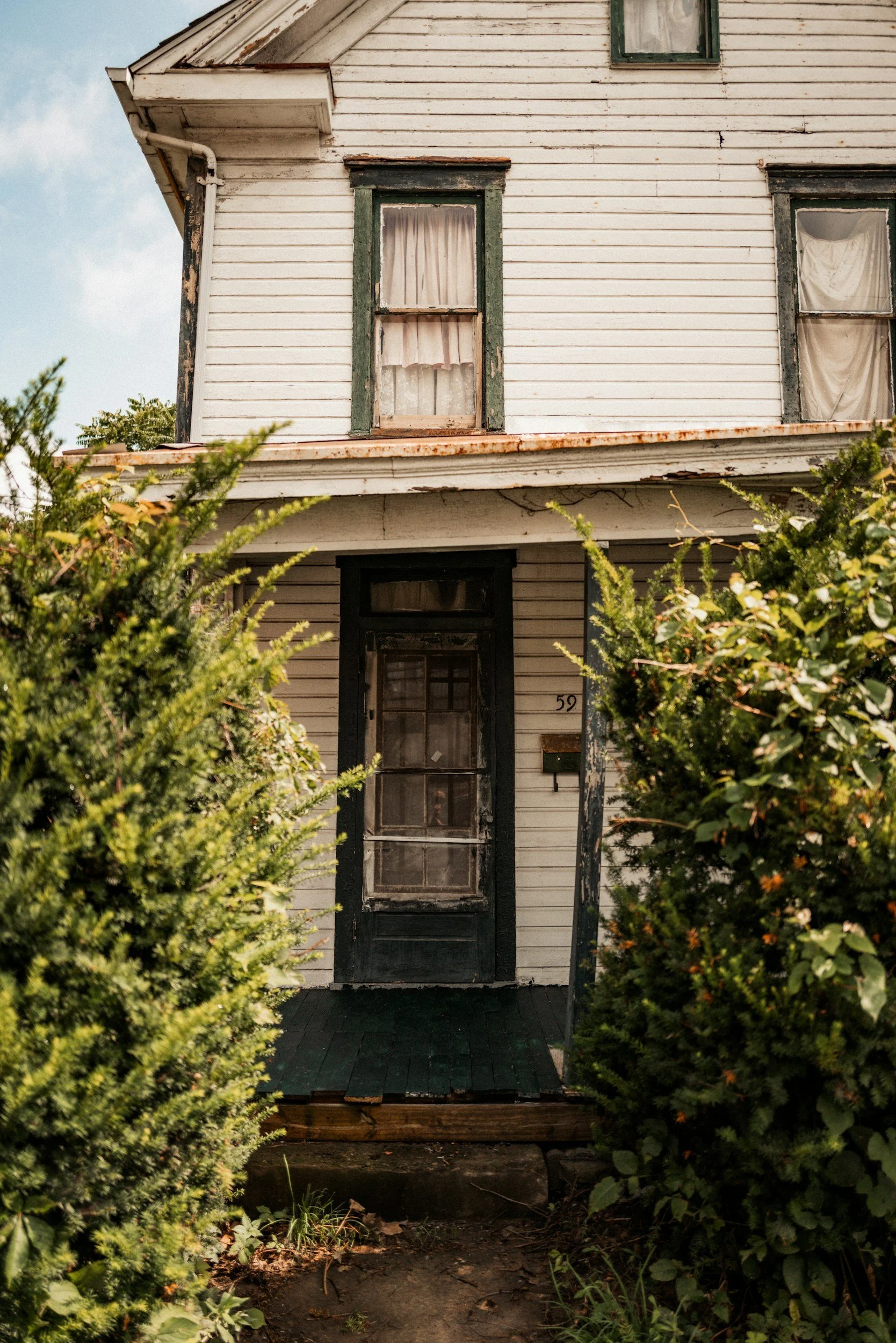 Old, weathered house with peeling paint, boarded-up windows, and a dark door, surrounded by overgrown bushes and plants.