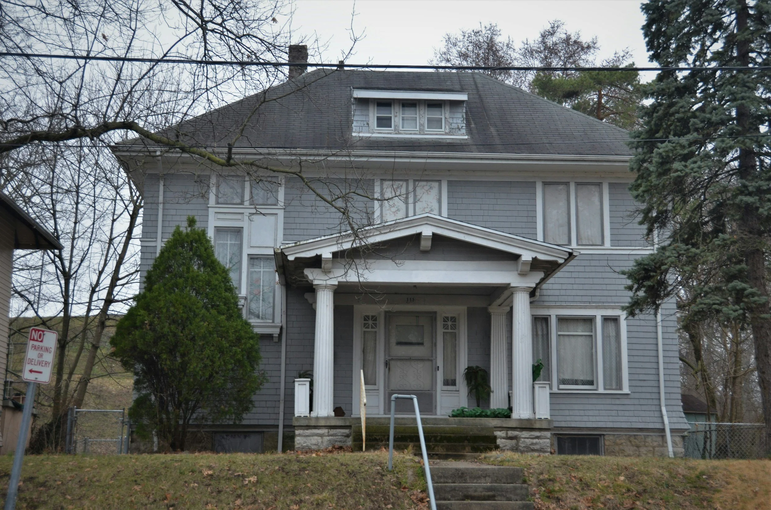 Front view of a large, old, gray wooden house with a covered porch, steps leading to the entrance, and a dormer window on the roof. There are leafless trees and greenery in the yard, and a