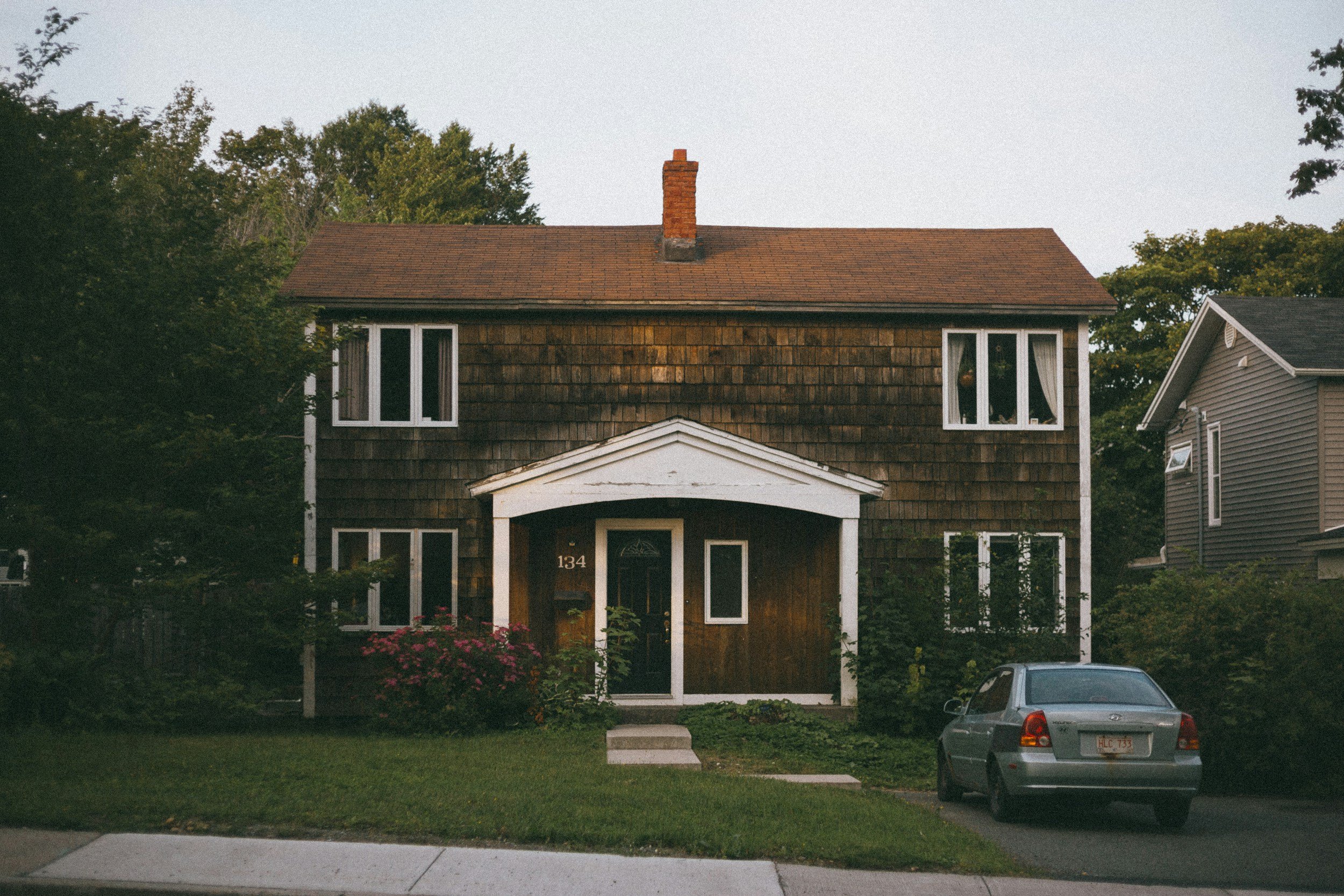 A two-story house with brown wooden shingles on the upper floor, white trim, and a brick chimney. There is a porch with a white triangular pediment, a black door, and windows with white framing. The house number '134' is visible near the door. The house is surrounded by greenery, including bushes and trees. A gray car is parked in front of the house on a driveway, and the sky is clear.
